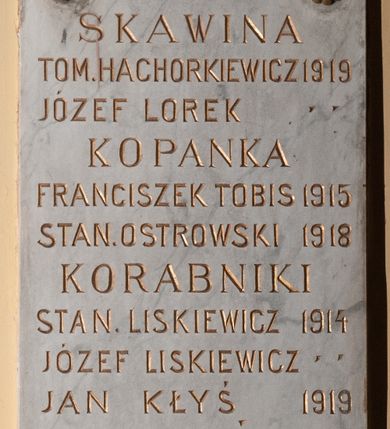 Zdjęcie nr 1: Tablica pamiątkowa z jasnego kamienia w kształcie bardzo wydłużonego, stojącego prostokąta, z kutym, złoconym napisem „SKAWINA / TOM(ASZ) HACHORKIEWICZ 1919 / JÓZEF LOREK / KOPANKA / FRANCISZEK TOBIS 1915 / STAI(ISŁAW) OSTROWSKI 1918 / KORABNIKI / STAN(ISŁAW) LISKIEWICZ 1914 / JÓZEF LISKIEWICZ / JAN KŁYŚ 1919 / RZOZÓW / JÓZEF PLANTA 1914 / JAN KUBAS 1915 / MICHAŁ MADEJ / ANTONI KLIMAS 1918 / MICHAŁ PUTAJ / SAMBOREK / PIOTR HABAS 1917 / SIDZINA / JAN STYRYLSKI 1914 / ANDRZEJ BYK / JÓZEF CHMURA / MICHAŁ BALON 1915 / FRANCISZEK BYK 1919”. W narożach ozdobne rozetki maskują miejsca mocowania.
