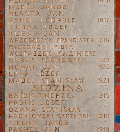 Zdjęcie nr 1: Tablica pamiątkowa z jasnego kamienia w kształcie bardzo wydłużonego, stojącego prostokąta, z kutym, złoconym napisem „RZOZÓW / PŁONKA JÓZEF 1914 / OSTROGORSKI JAN / KACZMARA JAN  1915 / MADEJ MICHAŁ / PANEK ANDRZEJ / JANIK JÓZEF / MADEJ STANISŁAW / BALON STEFAN / BALON MICHAŁ / PŁONKA PIOTR 1916 / MADEJ JAKÓB / PLANTA JAN / PANEK LEOPOLD 1917 / KUBAS JÓZEF / KUBAS JAN / WRZOSZCZYK FRANCISZEK 1918 / RYDZOWSKI PIOTR / WOJTASZEK KAZIMIERZ / KUBAS FRANCISZEK 1919 / NOWAK JAN 1920 / LUPA JÓZEF / MADEJ STANISŁAW 1921 / SIDZINA / BALON ANDRZEJ 1915 / PROFIC JÓZEF / OZANA STANISŁAW / HACHLOWSKI SZCZEPAN 1916 / ZIELNIK JAKÓB / PACHEL JAN 1918 / PACHEL JÓZEF / WRONA WOJCIECH / PACHEL LUDWIK 1920 / KOPANKA / OSTROWSKI JAN 1914 / KLIMAS JAN 1915 / TOBIS HENRYK / FAJTO FRANCISZEK 1916 / HAJTO WŁADYSŁAW / ZAPOMNIANEMU / ŻOŁNIERZOWI”. W narożach ozdobne rozetki maskują miejsca mocowania.