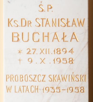 Zdjęcie nr 1: Tablica z jasnego kamienia w kształcie bardzo wydłużonego, stojącego prostokąta, z kutym, złoconym napisem „Ś(WIĘTEJ) P(AMIĘCI) / KS(IĄDZ) D[OKTO]R STANISŁAW / BUCHAŁA / *27. XII. 1894 / + 9. X. 1958 / PROBOSZCZ SKAWIŃSKI / W LATACH 1935-1958 / PROPAGATOR / AKCJI KATOLICKIEJ / I CHRZEŚCIJAŃSKICH / ZWIĄZKÓW ZAWODOWYCH / WIĘZIEŃ CZASÓW / STALINOWSKICH / BEZ RESZTY ODDANY BOGU / OJCZYZNIE I KOŚIOŁOWI / W 50-TĄ ROCZNICĘ ŚMIERCI / MIESZKAŃCY SKAWINY”. W narożach ozdobne rozetki maskują miejsca mocowania epitafium. 