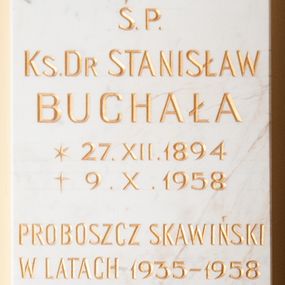 Zdjęcie nr 1: Tablica z jasnego kamienia w kształcie bardzo wydłużonego, stojącego prostokąta, z kutym, złoconym napisem „Ś(WIĘTEJ) P(AMIĘCI) / KS(IĄDZ) D[OKTO]R STANISŁAW / BUCHAŁA / *27. XII. 1894 / + 9. X. 1958 / PROBOSZCZ SKAWIŃSKI / W LATACH 1935-1958 / PROPAGATOR / AKCJI KATOLICKIEJ / I CHRZEŚCIJAŃSKICH / ZWIĄZKÓW ZAWODOWYCH / WIĘZIEŃ CZASÓW / STALINOWSKICH / BEZ RESZTY ODDANY BOGU / OJCZYZNIE I KOŚIOŁOWI / W 50-TĄ ROCZNICĘ ŚMIERCI / MIESZKAŃCY SKAWINY”. W narożach ozdobne rozetki maskują miejsca mocowania epitafium. 