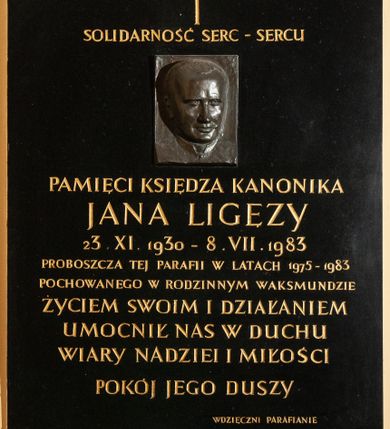 Zdjęcie nr 1: Epitafium w kształcie stojącego prostokąta wykonane z czarnego kamienia poświęcone pamięci księdza Jana Ligęzy. Na tablicy poniżej krzyża łacińskiego i napisu „SOLIDARNOŚĆ SERC – SERCU” płaskorzeźbiony portret zmarłego. Na owalnej twarzy z wysokim czołem odznaczają się małe oczy, wąski nos i wydatne usta, między nosem a ustami zaznaczona bruzda. Pod portretem dalsza część napisu „/ PAMIĘCI KSIĘDZA KANONIKA / JANA LIGĘZY / 23. IX. 1930 – 8. VII. 1983 / PROBOSZCZA TEJ PARAFII W LATACH 1975-1983 / POCHOWANEGO W RODZINNYM WAKSMUNDZIE / ŻYCIEM SWOIM I DZIAŁANIEM / UMOCNIŁ NAS W DUCHU / WIARY NADZIEI I MIŁOŚCI / POKÓJ JEGO DUSZY / WDZIĘCZNI PARAFIANIE”. 