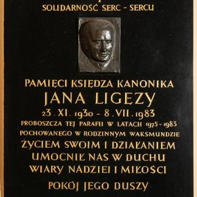Zdjęcie nr 1: Epitafium w kształcie stojącego prostokąta wykonane z czarnego kamienia poświęcone pamięci księdza Jana Ligęzy. Na tablicy poniżej krzyża łacińskiego i napisu „SOLIDARNOŚĆ SERC – SERCU” płaskorzeźbiony portret zmarłego. Na owalnej twarzy z wysokim czołem odznaczają się małe oczy, wąski nos i wydatne usta, między nosem a ustami zaznaczona bruzda. Pod portretem dalsza część napisu „/ PAMIĘCI KSIĘDZA KANONIKA / JANA LIGĘZY / 23. IX. 1930 – 8. VII. 1983 / PROBOSZCZA TEJ PARAFII W LATACH 1975-1983 / POCHOWANEGO W RODZINNYM WAKSMUNDZIE / ŻYCIEM SWOIM I DZIAŁANIEM / UMOCNIŁ NAS W DUCHU / WIARY NADZIEI I MIŁOŚCI / POKÓJ JEGO DUSZY / WDZIĘCZNI PARAFIANIE”. 