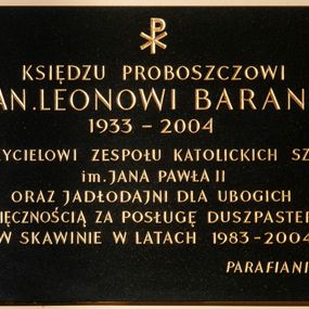 Zdjęcie nr 1: Tablica w kształcie leżącego prostokąta z kutym, złoconym napisem poniżej chrystogramu „KSIĘDZU PROBOSZCZOWI / KS(IĘDZU) KAN(ONIKOWI) LEONOWI BARANOWI / 1933-2004 / ZAŁOŻYCIELOWI ZESPOŁU KATOLICKICH SZKÓŁ / im(ienia) JANA PAWŁA II / ORAZ JADŁODAJNI DLA UBOGICH / Z WDZIĘCZNOŚCIĄ ZA POSŁUGĘ DUSZPASTERSKĄ / W SKAWINIE W LATACH 1983-2004 / PARAFIANIE”. W narożach dekoracyjne rozetki maskują miejsca mocowania. 