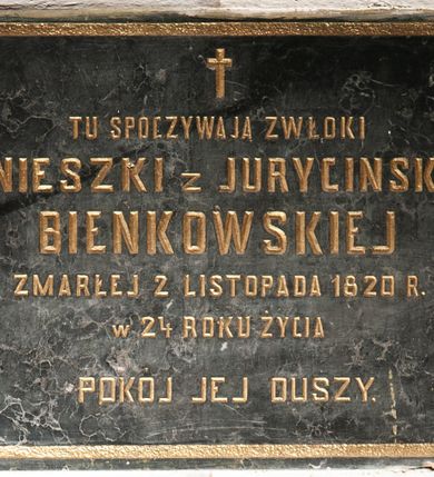 Zdjęcie nr 1: Tablica w kształcie leżącego prostokąta obwiedziona ramką z kwadratami w narożach. Na środku, poniżej krzyża łacińskiego złocona, majuskułowa inskrypcja „TU SPOCZYWAJĄ ZWŁOKI / AGNIESZKI z JURYCINSKICH / BIEŃKOWSKIEJ / ZMARŁEJ 2 LISTOPADA 1820 R(OKU) / W 24 ROKU ŻYCIA / POKÓJ JEJ DUSZY”. 