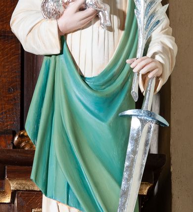 Zdjęcie nr 2: Figura pełna na cokole przedstawiająca św. Agnieszkę, która stoi frontalnie, kierując wzrok lekko w prawo. Ma owalną twarz, małe oczy, szeroki nos i małe usta oraz długie, falowane włosy, które opadają na ramiona i plecy; na głowie wianek z lilii. Prawą, uniesioną ręką przytrzymuje baranka, wyciągającego pyszczek w stronę liścia palmowego, który święta, razem z mieczem trzyma w lewej dłoni. Jest ubrana w białą suknię marszczoną pod szyją oraz zielony płaszcz przechodzący przez lewy bark i przytrzymany prawą ręką ponad biodrem. Polichromia w partiach ciała naturalistyczna.