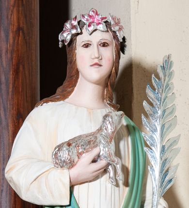 Zdjęcie nr 1: Figura pełna na cokole przedstawiająca św. Agnieszkę, która stoi frontalnie, kierując wzrok lekko w prawo. Ma owalną twarz, małe oczy, szeroki nos i małe usta oraz długie, falowane włosy, które opadają na ramiona i plecy; na głowie wianek z lilii. Prawą, uniesioną ręką przytrzymuje baranka, wyciągającego pyszczek w stronę liścia palmowego, który święta, razem z mieczem trzyma w lewej dłoni. Jest ubrana w białą suknię marszczoną pod szyją oraz zielony płaszcz przechodzący przez lewy bark i przytrzymany prawą ręką ponad biodrem. Polichromia w partiach ciała naturalistyczna.