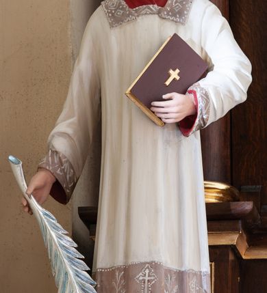 Zdjęcie nr 1: Figura pełna na cokole przedstawiająca św. Szczepana. Święty stoi frontalnie, kierując wzrok przed siebie. Ma owalną twarz, małe oczy, szeroki nos i usta o kącikach skierowanych w dół oraz krótkie włosy zaczesane na bok, z przedziałkiem. Prawą, opuszczoną ręką przytrzymuje gałązkę palmową, lewą przyciska do ciała zamkniętą księgę; u stóp dwa białe kamienie. Ubrany jest w białą rokietę zdobioną przy dolnej krawędzi, przy rękawach i pod szyją szerokim pasem koronki założoną na czerwoną tunikę. Polichromia w partiach ciała naturalistyczna. 