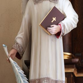 Zdjęcie nr 1: Figura pełna na cokole przedstawiająca św. Szczepana. Święty stoi frontalnie, kierując wzrok przed siebie. Ma owalną twarz, małe oczy, szeroki nos i usta o kącikach skierowanych w dół oraz krótkie włosy zaczesane na bok, z przedziałkiem. Prawą, opuszczoną ręką przytrzymuje gałązkę palmową, lewą przyciska do ciała zamkniętą księgę; u stóp dwa białe kamienie. Ubrany jest w białą rokietę zdobioną przy dolnej krawędzi, przy rękawach i pod szyją szerokim pasem koronki założoną na czerwoną tunikę. Polichromia w partiach ciała naturalistyczna. 