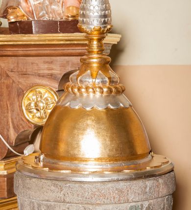 Zdjęcie nr 3: Chrzcielnica kamienna o formie kielichowej, składająca się z czary z pokrywą, trzonu oraz stopy. Stopa okrągłą, stanowiąca pierwotnie część żaren, w formie walca z otworem przy brzegu. Na niej zwężający się trzon złożony z nodusa i pierścieni. Na nodusie ornament plecionkowy, na pierścieniu lancetowate liście. Czara półokrągła, pokryta dekoracją z liści akantu, z gładką krawędzią i wydrążonym zagłębieniem na misę. Pokrywa drewniana, wysoka, półkolista, z szerokim otokiem dekorowanym półkolami; zwęża się ku górze. Rozbudowane zwieńczenie dekorowane od dołu kolejno koronką zakończoną półkoliście, puklowaniem, motywem płomieni oraz profilowanym trzonem zakończonym dużą szyszką. Chrzcielnica w kolorze kamienia, gdzieniegdzie pozostałości czerwonej farby, pokrywa złocona i srebrzona. 