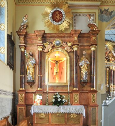 Zdjęcie nr 1: Nastawa ołtarzowa architektoniczna, trójosiowa, jednokondygnacyjna ze zwieńczeniem, na cokole. Mensa skrzyniowa, z antepedium artykułowanym trzema prostokątnymi płycinami, w bocznych rozety, w środkowej krzyż równoramienny. Cokół nastawy siedmiodzielny, z nieparzystymi częściami wysuniętymi przed lico, zdobionymi plakietami ornamentalnymi. Nastawa dzielona kolumnami z opaskami w jednej trzeciej wysokości, o kapitelach utworzonych z elementów roślinnych, podtrzymujących belkowanie wyłamane na ich osiach. W wyłamanych częściach fryzu plakiety dekoracyjne z motywem roślinnym. W polu centralnym nisza z rzeźbą ukrzyżowanego Chrystusa między narzędziami męki, oprawiona w szeroką, profilowaną ramę. Powyżej niej para putt trzymająca kartusz obwiedziony ramą z kwitnących róż z napisem „JEZU / ZMIŁUJ SIĘ / NAD / NAMI”. W polach bocznych ustawione na konsolach i cokołach figury św. Józefa po lewej i św. Joachima (?) po prawej stronie. W partii zwieńczenia urwane fragmenty przyczółka ponad zewnętrznymi fragmentami belkowania, na nich posadowione modlące się postacie. Na środku, w okrągłym polu monogram IHS otoczony obłokami, na tle glorii promienistej. 
Ołtarz marmoryzowany na kolor jasnobrązowy, płyciny na kolor brązowo-żółty, kolumny na ciemnobrązowy. Detale i ornamenty złocone; polichromia w partiach ciała postaci naturalistyczna. 