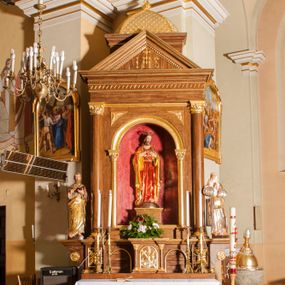 Zdjęcie nr 1: Ołtarz przyścienny, architektoniczny, jednoosiowy, jednokondygnacyjny. Mensa skrzyniowa z kamiennym portatylem, poprzedzona dwoma, masywnymi kolumienkami o kostkowych kapitelach. Antepedium artykułowane czterema prostokątnymi płycinami zamkniętymi półkoliście. Na mensie, na kolumienkach blat ołtarza. Na nim prostopadłościenne tabernakulum, z drzwiczkami akcentowanymi płyciną w kształcie prostokąta zamkniętego półkoliście. Na drzwiach aplikowane, złączone ze sobą symbole: kielich z hostią, gorejące serce, krzyż i kotwica na tle obłoków. Cokół nastawy ołtarza pięciodzielny, z płycinami. Wysunięte osie cokołu są dekorowane plakietą z motywem roślinnym, zaś skrajne są ozdobione konsolami z aplikowaną, okrągłą rozetą. Pole środkowe z prostokątną wnęką zamkniętą półkoliście, z łukiem wspartym na dwóch kolumnach korynckich. We wnęce na czerwonym tle figura Najświętszego Serca Pana Jezusa. W narożach plakiety ornamentalne w trójkątnych polach. Nastawa flankowana parą kolumn korynckich ustawionych na wysuniętych partiach cokołu oraz figurami świętych. Kolumny wspierają belkowanie zwieńczone trójkątnym przyczółkiem wypełnionym chrystogramem. Za tympanonem pozłacana kopuła pokryta motywem rybiej łuski, zwieńczona krzyżem postawionym na rozecie. Całość ołtarza fladrowana w odcieniu ciemnobrązowym, ornamenty i detal architektoniczny złocony, polichromia rzeźb naturalistyczna.
