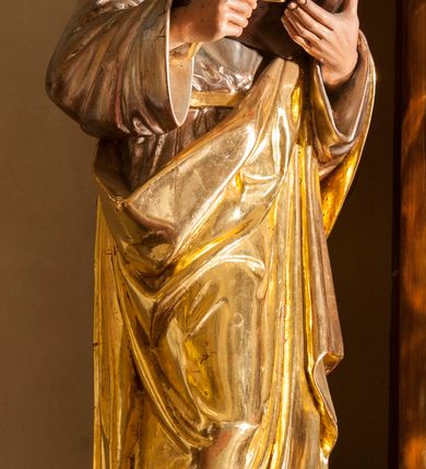 Zdjęcie nr 1: Figura pełna na cokole przedstawiająca mężczyznę. Święty stoi frontalnie, kierując wzrok przed siebie. Ma owalną twarz, migdałowe oczy, wąski nos i małe, grube usta oraz zakrywające uszy włosy zaczesane do tyłu, z lokiem nad czołem. W prawej, uniesionej ręce trzyma liść palmy lub pióro, lewą podtrzymuje otwartą księgę. Ubrany jest w srebrną tunikę przewiązaną złotym sznurem oraz złoty płaszcz, który okrywa lewą rękę i zawija się wokół prawego biodra, opadając w dół sztywnymi fałdami. Spod stroju wystają bose stopy. Polichromia w partiach ciała naturalistyczna.
