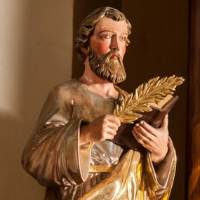 Zdjęcie nr 1: Figura pełna na cokole przedstawiająca mężczyznę. Święty stoi frontalnie, kierując wzrok przed siebie. Ma owalną twarz, migdałowe oczy, wąski nos i małe, grube usta oraz zakrywające uszy włosy zaczesane do tyłu, z lokiem nad czołem. W prawej, uniesionej ręce trzyma liść palmy lub pióro, lewą podtrzymuje otwartą księgę. Ubrany jest w srebrną tunikę przewiązaną złotym sznurem oraz złoty płaszcz, który okrywa lewą rękę i zawija się wokół prawego biodra, opadając w dół sztywnymi fałdami. Spod stroju wystają bose stopy. Polichromia w partiach ciała naturalistyczna.