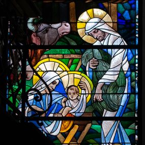 Zdjęcie nr 1: Witraż w kształcie stojącego prostokąta zamkniętego łukiem półkolistym. W centrum kompozycji, na tle stajenki ukazana Święta Rodzina. Z lewej strony znajduje się przedstawiona z profilu, w pozycji klęczącej Maria pochylająca się nad leżącym w żłóbku Dzieciątkiem Jezus. Ukazana jest jako młoda kobieta, ubrana w niebiesko-białe szaty z białą chustą na głowie. W dłoniach podtrzymuje fragment tkaniny, którą otula Jezusa. Dzieciątko zostało przedstawione frontalnie, w pozycji siedzącej w drewnianym żłóbku o skrzyżowanych nogach. Obie rączki Jezusa ugięte w łokciach i uniesione na wysokość twarzy. Bose stopy wyciągnięte do przodu. Wokół głów Marii i Dzieciątka znajdują się świetliste nimby. Z prawej strony kompozycji przedstawiono św. Józefa ukazanego w pozycji stojącej, z ciałem skierowanym w trzech czwartych w prawo. Święty pochyla się nieco do przodu w kierunku żłóbka. Ma twarz dojrzałego mężczyzny, z krótką, brązową brodą. W ręku trzyma fragment futrzanego okrycia. Ubrany jest w biało-niebieskie, długie szaty oraz białą chustę. Stopy postaci są bose. Wokół głowy świętego znajduje się świetlisty nimb. Na drugim planie, z prawej strony przedstawiono zwierzęta: wołu oraz osła i drewnianą stajenkę, a w tle rozgwieżdżone, granatowe niebo z widoczną gwiazdą betlejemską. W dolnej części kompozycji, na pierwszym planie ukazane są owce i barany. W prawym dolnym rogu witraża umieszczona jest sygnatura „Adam Bunsch / 1960”.