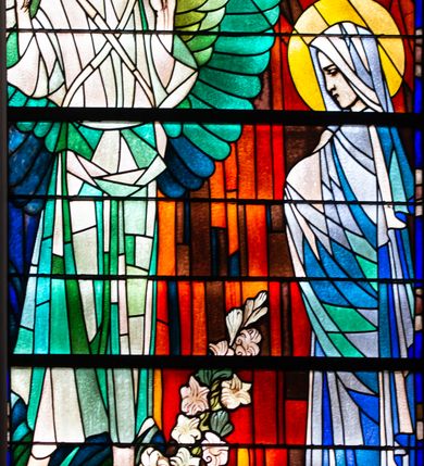 Zdjęcie nr 2: Witraż w kształcie stojącego prostokąta zamkniętego łukiem półkolistym. W centrum kompozycji, na tle czerwonej kotary ukazana scena Zwiastowania. Z prawej strony znajduje się przedstawiona z profilu, w pozycji stojącej Maria ubrana w niebiesko-białe szaty. Ukazana jest jako młoda kobieta z pochyloną głową. Ręce schowane pod połami szat, złożone na piersi. Na głowie Marii jasnoniebieska chusta, wokół niej świetlisty nimb. Z prawej strony kompozycji przedstawiono archanioła Gabriela ukazanego frontalnie, w pozycji stojącej. Głowa pochylona w trzech czwartych w prawo. Archanioł ma pozbawioną zarostu twarz młodego mężczyzny i brązowe, sięgające uszu włosy. Ubrany jest w biało-zielone szaty i żółte sandały. Obie ręce, ugięte w łokciach, uniesione w geście pozdrowienia. Szeroko rozpostarte skrzydła archanioła mają pióra w odcieniach zieleni i niebieskiego. Wokół głowy postaci znajduje się niebiesko-zielony nimb. W górnej części kompozycji ukazana gołębica Ducha Świętego na tle promienia skierowanego na głowię Marii. Na drugim planie granatowe niebo. W dolnej części kompozycji, na pierwszym planie ukazane są białe lilie.