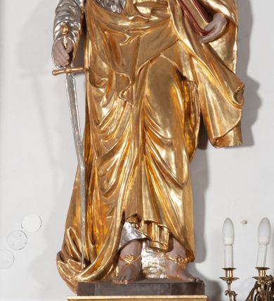 Zdjęcie nr 1: Całopostaciowa, ustawiona na konsoli w formie spływu wolutowego figura przedstawia zwróconą na wprost postać św. Pawła stojącego w lekkim kontrapoście. Pociągłą twarz charakteryzuje długi, ciemnobrązowy zarost, niewielkie, szeroko otwarte oczy wzniesione brwi oraz długi, prosty nos. Włosy są krótkie, ciemnobrązowe, kręcone. Święty ubrany jest w srebrzoną suknię z długimi rękawami oraz osłaniający niemal całą sylwetkę, wydatnie fałdowany złocony płaszcz, na stopach są złocone sandały. Prawą, opuszczoną w dół ręką podtrzymuje wsparty na pole płaszcza miecz, w lewej ma księgę w czerwonej oprawie. 