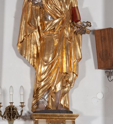 Zdjęcie nr 1: Całopostaciowa, ustawiona na konsoli w formie spływu wolutowego figura przedstawia zwróconą na wprost postać św. Piotra stojącego w lekkim kontrapoście. Owalną twarz charakteryzuje krótki, ciemnobrązowy zarost, niewielkie, szeroko otwarte oczy wzniesione brwi oraz długi, prosty nos. Włosy są krótkie, ciemnobrązowe, kręcone. Święty ubrany jest w srebrzoną suknię z długimi rękawami oraz osłaniający niemal całą sylwetkę, wydatnie fałdowany złocony płaszcz. Prawą, zgiętą w łokciu rękę wznosi do góry w geście błogosławieństwa. Lewą podtrzymuje księgę w czerwonej oprawie, w dłoni ma parę skrzyżowanych, złoconych kluczy.  