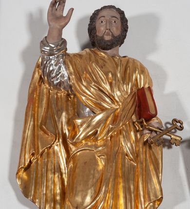 Zdjęcie nr 2: Całopostaciowa, ustawiona na konsoli w formie spływu wolutowego figura przedstawia zwróconą na wprost postać św. Piotra stojącego w lekkim kontrapoście. Owalną twarz charakteryzuje krótki, ciemnobrązowy zarost, niewielkie, szeroko otwarte oczy wzniesione brwi oraz długi, prosty nos. Włosy są krótkie, ciemnobrązowe, kręcone. Święty ubrany jest w srebrzoną suknię z długimi rękawami oraz osłaniający niemal całą sylwetkę, wydatnie fałdowany złocony płaszcz. Prawą, zgiętą w łokciu rękę wznosi do góry w geście błogosławieństwa. Lewą podtrzymuje księgę w czerwonej oprawie, w dłoni ma parę skrzyżowanych, złoconych kluczy.  
