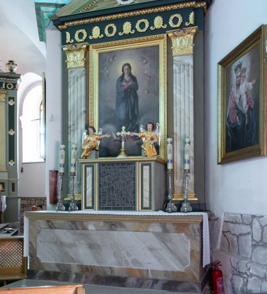 Zdjęcie nr 2: Nastawa architektoniczna, jednoosiowa, jednokondygnacyjna. Ołtarz drewniany, prostopadłościenny, na cokole, z malowaną płyciną na ściance przedniej w formie leżącego prostokąta o wklęsłych bokach, polichromowane na sposób imitujący marmoryzację w różnych odcieniach brązu. Zakończone profilowanym gzymsem, dekorowanym złoconym ornamentem roślinnym. Mensa drewniana. Na ołtarzu ustawione tabernakulum w formie drewnianej, prostopadłościennej skrzynki. Na przedniej ściance witraż z przedstawieniem sceny Ukrzyżowania, flankowany dwiema płycinami. Całość obwiedziona profilowanym, złoconym gzymsem. Na tabernakulum dwa cokoły z siedzącymi aniołami. Nastawa ujęta dwoma pilastrami o kanelowanych trzonach, dźwigających niepełne belkowanie i trójkątny przyczółek. Fryz dekorowany ustawionymi naprzemiennie anthemionami i wieńcami, belkowanie i przyczółek zdobione kimationem. W polu przyczółka owalny medalion z monogramem Marii, ujęty wieńcem, w glorii promienistej. W polu środkowym obraz w formie stojącego prostokąta z wizerunkiem Matki Boskiej, ujęty profilowaną ramą dekorowaną kimationem. Nastawa polichromowana jest w kolorze szarym, trzony pilastrów są marmoryzowane, belkowanie o barwie zielono-niebieskiej. Profile, kapitele i dekoracja złocone, wieniec srebrzony. 