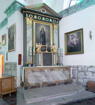 Zdjęcie nr 3: Nastawa architektoniczna, jednoosiowa, jednokondygnacyjna. Ołtarz drewniany, prostopadłościenny, na cokole, z malowaną płyciną na ściance przedniej w formie leżącego prostokąta o wklęsłych bokach, polichromowane na sposób imitujący marmoryzację w różnych odcieniach brązu. Zakończone profilowanym gzymsem, dekorowanym złoconym ornamentem roślinnym. Mensa drewniana. Na ołtarzu ustawione tabernakulum w formie drewnianej, prostopadłościennej skrzynki. Na przedniej ściance witraż z przedstawieniem sceny Ukrzyżowania, flankowany dwiema płycinami. Całość obwiedziona profilowanym, złoconym gzymsem. Na tabernakulum dwa cokoły z siedzącymi aniołami. Nastawa ujęta dwoma pilastrami o kanelowanych trzonach, dźwigających niepełne belkowanie i trójkątny przyczółek. Fryz dekorowany ustawionymi naprzemiennie anthemionami i wieńcami, belkowanie i przyczółek zdobione kimationem. W polu przyczółka owalny medalion z monogramem Marii, ujęty wieńcem, w glorii promienistej. W polu środkowym obraz w formie stojącego prostokąta z wizerunkiem Matki Boskiej, ujęty profilowaną ramą dekorowaną kimationem. Nastawa polichromowana jest w kolorze szarym, trzony pilastrów są marmoryzowane, belkowanie o barwie zielono-niebieskiej. Profile, kapitele i dekoracja złocone, wieniec srebrzony. 