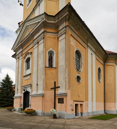 Zdjęcie nr 16: Kościół jest orientowany, murowany z cegły i kamienia, na rzucie podłużnym. Zajmuje powierzchnię około 606 metrów kwadratowych, długość bryły wynosi około 32, a szerokość 14 metrów. Jednonawowy, trójprzęsłowy korpus charakteryzuje środkowe, kwadratowe przęsło z płytkimi apsydami po bokach, wysuwającymi się przed lica elewacji bocznych, na wzór pseudotranseptu (para spłaszczonych apsyd). Przęsło wschodnie, na rzucie prostokąta, z wnękami po bokach, zakończone jest apsydą stanowiącą przestrzeń prezbiterium. Na osi kościoła i przedłużeniu prezbiterium jest piętrowa zakrystia ze skarbcem, natomiast w przedłużeniu przęsła zachodniego wieża ujęta po bokach piętrowymi lokalnościami. Korpus nawowy przekryty jest sklepieniem kolebkowym na gurtach, z lunetami, które w przęśle środkowym, na przecięciu z pseudotranseptem, tworzy rodzaj sklepienia krzyżowego z okrągłym zwierciadłem. Artykulację ścian wewnątrz kościoła stanowią pary pilastrów wspierające przerywane belkowanie. Na zewnątrz ściany korpusu, transeptu i zakrystii artykułują osadzone na niewysokim cokole lizeny wspierające obiegające całość belkowanie. Dodatkowo mury przeprute są owalnymi otworami okiennymi ujętymi profilowanymi obramieniami. Fasadę charakteryzuje w części środkowej ryzalit o skośnych ściankach bocznych oraz podziały podwójnymi i pojedynczymi pilastrami jońskimi, osadzonymi na wysokich cokołach i wspierającymi belkowanie. Do wnętrza kościoła prowadzą trzy otwory drzwiowe – w elewacji południowej, północnej i zachodniej. Drzwi w fasadzie są ujęte dekoracyjnym portalem z parą wąskich pilastrów, zwieńczonych zwężającymi się ku dołowi kapitelami, wspierających gzyms w formie łuku odcinkowego. Całość ujmuje obramienie o fantazyjnym wykroju w górnej partii, którego krawędzie obiega profilowana listwa z parą niewielkich wolut w górnych narożach. Obramienie drzwi wieńczy prosty gzyms odcinkowy nakryty blaszanym daszkiem. Po bokach wejścia znajduje się para kamiennych kropielnic. Powyżej otworu drzwiowego, pole środkowe fasady przeprute zostało oknem w kształcie stojącego prostokąta zamkniętego łukiem odcinkowym, zamurowanego w jednej trzeciej wysokości. Obramienie okna jest w formie pary pilastrów bez baz, wspierających fryz z kluczem pośrodku i parą wsporników wolutowych po bokach, dźwigających odcinkowy gzyms wieńczący. Pod parapetem umieszczono fartuch w kształcie leżącego prostokąta, o wklęsłych dolnych narożach, z dolną krawędzią zdobioną motywem lambrekinu. W skośnych ścianach bocznych ryzalitu znajduje się para zamkniętych półkoliście niszy z figurami św. Piotra po lewej i św. Pawła po prawej stronie. 
Ryzalit wieńczy trójkątny przyczółek z herbem Nowina w tympanonie. Nad belkowaniem, na całej szerokości fasady, znajduje się prosta attyka z odpowiadającymi dolnej strefie elewacji podziałami lizenowymi, wspierającymi wyłamujący się ponad nimi gzyms ozdobiony wazonami. Powyżej wznosi się górne piętro wieży, czworościenne o ściętych narożnikach, nakryte kopulastym hełmem z nasadnikiem.  
Północna, zachodnia i południowa ściana wieży przeprute są otworami okiennymi w kształcie stojącego prostokąta zamkniętego łukiem odcinkowym. Okna mają obramienia w kształcie stojących prostokątów z uszakami w górnych narożach, zwieńczone są belkowaniem w formie łuku odcinkowego z kluczem pośrodku i parą wolutowych wsporników po bokach fryzu. Podwieszenie ma formę fartucha w kształcie leżącego prostokąta o wklęsłych dolnych narożach i dolnej krawędzi zdobionej motywem lambrekinu. Powyżej otworów okiennych znajdują się okrągłe tarcze zegarowe. Pola ściętych naroży wieży zdobi ornamentyka cekinowa i kampanule. Korpus nawowy przykrywa dwuspadowy dach zakończony pięciopołaciowo nad prezbiterium i trójpołaciowo nad końcami transeptu, kryty dachówką. Na skrzyżowaniu korpusu nawowego i transeptu ulokowano obitą blachą wieżyczkę na sygnaturkę. Kolorystyka tynków kościoła utrzymana jest w odcieniu ugru, w dolnej partii różu, detal architektoniczny i ornamentyka w bieli. 

Plac przykościelny otoczony jest murowanym parkanem z usytuowanymi w nim 14 kapliczkami ze stacjami drogi krzyżowej, nakrytymi dwuspadowymi dachami. W jednym z naroży znajduje się niewielka, murowana kaplica grobowa rodziny Romerów o ceglanych ścianach artykułowanych białymi pilastrami i fasadzie ze szczytem ujętym parą spływów i zwieńczonym trójkątnym przyczółkiem.   
