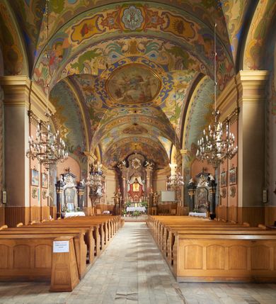 Zdjęcie nr 35: Kościół jest orientowany, murowany z cegły i kamienia, na rzucie podłużnym. Zajmuje powierzchnię około 606 metrów kwadratowych, długość bryły wynosi około 32, a szerokość 14 metrów. Jednonawowy, trójprzęsłowy korpus charakteryzuje środkowe, kwadratowe przęsło z płytkimi apsydami po bokach, wysuwającymi się przed lica elewacji bocznych, na wzór pseudotranseptu (para spłaszczonych apsyd). Przęsło wschodnie, na rzucie prostokąta, z wnękami po bokach, zakończone jest apsydą stanowiącą przestrzeń prezbiterium. Na osi kościoła i przedłużeniu prezbiterium jest piętrowa zakrystia ze skarbcem, natomiast w przedłużeniu przęsła zachodniego wieża ujęta po bokach piętrowymi lokalnościami. Korpus nawowy przekryty jest sklepieniem kolebkowym na gurtach, z lunetami, które w przęśle środkowym, na przecięciu z pseudotranseptem, tworzy rodzaj sklepienia krzyżowego z okrągłym zwierciadłem. Artykulację ścian wewnątrz kościoła stanowią pary pilastrów wspierające przerywane belkowanie. Na zewnątrz ściany korpusu, transeptu i zakrystii artykułują osadzone na niewysokim cokole lizeny wspierające obiegające całość belkowanie. Dodatkowo mury przeprute są owalnymi otworami okiennymi ujętymi profilowanymi obramieniami. Fasadę charakteryzuje w części środkowej ryzalit o skośnych ściankach bocznych oraz podziały podwójnymi i pojedynczymi pilastrami jońskimi, osadzonymi na wysokich cokołach i wspierającymi belkowanie. Do wnętrza kościoła prowadzą trzy otwory drzwiowe – w elewacji południowej, północnej i zachodniej. Drzwi w fasadzie są ujęte dekoracyjnym portalem z parą wąskich pilastrów, zwieńczonych zwężającymi się ku dołowi kapitelami, wspierających gzyms w formie łuku odcinkowego. Całość ujmuje obramienie o fantazyjnym wykroju w górnej partii, którego krawędzie obiega profilowana listwa z parą niewielkich wolut w górnych narożach. Obramienie drzwi wieńczy prosty gzyms odcinkowy nakryty blaszanym daszkiem. Po bokach wejścia znajduje się para kamiennych kropielnic. Powyżej otworu drzwiowego, pole środkowe fasady przeprute zostało oknem w kształcie stojącego prostokąta zamkniętego łukiem odcinkowym, zamurowanego w jednej trzeciej wysokości. Obramienie okna jest w formie pary pilastrów bez baz, wspierających fryz z kluczem pośrodku i parą wsporników wolutowych po bokach, dźwigających odcinkowy gzyms wieńczący. Pod parapetem umieszczono fartuch w kształcie leżącego prostokąta, o wklęsłych dolnych narożach, z dolną krawędzią zdobioną motywem lambrekinu. W skośnych ścianach bocznych ryzalitu znajduje się para zamkniętych półkoliście niszy z figurami św. Piotra po lewej i św. Pawła po prawej stronie. 
Ryzalit wieńczy trójkątny przyczółek z herbem Nowina w tympanonie. Nad belkowaniem, na całej szerokości fasady, znajduje się prosta attyka z odpowiadającymi dolnej strefie elewacji podziałami lizenowymi, wspierającymi wyłamujący się ponad nimi gzyms ozdobiony wazonami. Powyżej wznosi się górne piętro wieży, czworościenne o ściętych narożnikach, nakryte kopulastym hełmem z nasadnikiem.  
Północna, zachodnia i południowa ściana wieży przeprute są otworami okiennymi w kształcie stojącego prostokąta zamkniętego łukiem odcinkowym. Okna mają obramienia w kształcie stojących prostokątów z uszakami w górnych narożach, zwieńczone są belkowaniem w formie łuku odcinkowego z kluczem pośrodku i parą wolutowych wsporników po bokach fryzu. Podwieszenie ma formę fartucha w kształcie leżącego prostokąta o wklęsłych dolnych narożach i dolnej krawędzi zdobionej motywem lambrekinu. Powyżej otworów okiennych znajdują się okrągłe tarcze zegarowe. Pola ściętych naroży wieży zdobi ornamentyka cekinowa i kampanule. Korpus nawowy przykrywa dwuspadowy dach zakończony pięciopołaciowo nad prezbiterium i trójpołaciowo nad końcami transeptu, kryty dachówką. Na skrzyżowaniu korpusu nawowego i transeptu ulokowano obitą blachą wieżyczkę na sygnaturkę. Kolorystyka tynków kościoła utrzymana jest w odcieniu ugru, w dolnej partii różu, detal architektoniczny i ornamentyka w bieli. 

Plac przykościelny otoczony jest murowanym parkanem z usytuowanymi w nim 14 kapliczkami ze stacjami drogi krzyżowej, nakrytymi dwuspadowymi dachami. W jednym z naroży znajduje się niewielka, murowana kaplica grobowa rodziny Romerów o ceglanych ścianach artykułowanych białymi pilastrami i fasadzie ze szczytem ujętym parą spływów i zwieńczonym trójkątnym przyczółkiem.   
