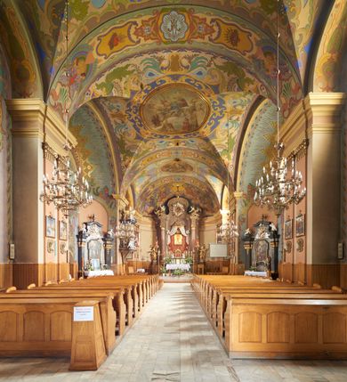 Zdjęcie nr 38: Kościół jest orientowany, murowany z cegły i kamienia, na rzucie podłużnym. Zajmuje powierzchnię około 606 metrów kwadratowych, długość bryły wynosi około 32, a szerokość 14 metrów. Jednonawowy, trójprzęsłowy korpus charakteryzuje środkowe, kwadratowe przęsło z płytkimi apsydami po bokach, wysuwającymi się przed lica elewacji bocznych, na wzór pseudotranseptu (para spłaszczonych apsyd). Przęsło wschodnie, na rzucie prostokąta, z wnękami po bokach, zakończone jest apsydą stanowiącą przestrzeń prezbiterium. Na osi kościoła i przedłużeniu prezbiterium jest piętrowa zakrystia ze skarbcem, natomiast w przedłużeniu przęsła zachodniego wieża ujęta po bokach piętrowymi lokalnościami. Korpus nawowy przekryty jest sklepieniem kolebkowym na gurtach, z lunetami, które w przęśle środkowym, na przecięciu z pseudotranseptem, tworzy rodzaj sklepienia krzyżowego z okrągłym zwierciadłem. Artykulację ścian wewnątrz kościoła stanowią pary pilastrów wspierające przerywane belkowanie. Na zewnątrz ściany korpusu, transeptu i zakrystii artykułują osadzone na niewysokim cokole lizeny wspierające obiegające całość belkowanie. Dodatkowo mury przeprute są owalnymi otworami okiennymi ujętymi profilowanymi obramieniami. Fasadę charakteryzuje w części środkowej ryzalit o skośnych ściankach bocznych oraz podziały podwójnymi i pojedynczymi pilastrami jońskimi, osadzonymi na wysokich cokołach i wspierającymi belkowanie. Do wnętrza kościoła prowadzą trzy otwory drzwiowe – w elewacji południowej, północnej i zachodniej. Drzwi w fasadzie są ujęte dekoracyjnym portalem z parą wąskich pilastrów, zwieńczonych zwężającymi się ku dołowi kapitelami, wspierających gzyms w formie łuku odcinkowego. Całość ujmuje obramienie o fantazyjnym wykroju w górnej partii, którego krawędzie obiega profilowana listwa z parą niewielkich wolut w górnych narożach. Obramienie drzwi wieńczy prosty gzyms odcinkowy nakryty blaszanym daszkiem. Po bokach wejścia znajduje się para kamiennych kropielnic. Powyżej otworu drzwiowego, pole środkowe fasady przeprute zostało oknem w kształcie stojącego prostokąta zamkniętego łukiem odcinkowym, zamurowanego w jednej trzeciej wysokości. Obramienie okna jest w formie pary pilastrów bez baz, wspierających fryz z kluczem pośrodku i parą wsporników wolutowych po bokach, dźwigających odcinkowy gzyms wieńczący. Pod parapetem umieszczono fartuch w kształcie leżącego prostokąta, o wklęsłych dolnych narożach, z dolną krawędzią zdobioną motywem lambrekinu. W skośnych ścianach bocznych ryzalitu znajduje się para zamkniętych półkoliście niszy z figurami św. Piotra po lewej i św. Pawła po prawej stronie. 
Ryzalit wieńczy trójkątny przyczółek z herbem Nowina w tympanonie. Nad belkowaniem, na całej szerokości fasady, znajduje się prosta attyka z odpowiadającymi dolnej strefie elewacji podziałami lizenowymi, wspierającymi wyłamujący się ponad nimi gzyms ozdobiony wazonami. Powyżej wznosi się górne piętro wieży, czworościenne o ściętych narożnikach, nakryte kopulastym hełmem z nasadnikiem.  
Północna, zachodnia i południowa ściana wieży przeprute są otworami okiennymi w kształcie stojącego prostokąta zamkniętego łukiem odcinkowym. Okna mają obramienia w kształcie stojących prostokątów z uszakami w górnych narożach, zwieńczone są belkowaniem w formie łuku odcinkowego z kluczem pośrodku i parą wolutowych wsporników po bokach fryzu. Podwieszenie ma formę fartucha w kształcie leżącego prostokąta o wklęsłych dolnych narożach i dolnej krawędzi zdobionej motywem lambrekinu. Powyżej otworów okiennych znajdują się okrągłe tarcze zegarowe. Pola ściętych naroży wieży zdobi ornamentyka cekinowa i kampanule. Korpus nawowy przykrywa dwuspadowy dach zakończony pięciopołaciowo nad prezbiterium i trójpołaciowo nad końcami transeptu, kryty dachówką. Na skrzyżowaniu korpusu nawowego i transeptu ulokowano obitą blachą wieżyczkę na sygnaturkę. Kolorystyka tynków kościoła utrzymana jest w odcieniu ugru, w dolnej partii różu, detal architektoniczny i ornamentyka w bieli. 

Plac przykościelny otoczony jest murowanym parkanem z usytuowanymi w nim 14 kapliczkami ze stacjami drogi krzyżowej, nakrytymi dwuspadowymi dachami. W jednym z naroży znajduje się niewielka, murowana kaplica grobowa rodziny Romerów o ceglanych ścianach artykułowanych białymi pilastrami i fasadzie ze szczytem ujętym parą spływów i zwieńczonym trójkątnym przyczółkiem.   
