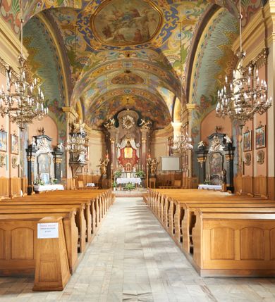 Zdjęcie nr 37: Kościół jest orientowany, murowany z cegły i kamienia, na rzucie podłużnym. Zajmuje powierzchnię około 606 metrów kwadratowych, długość bryły wynosi około 32, a szerokość 14 metrów. Jednonawowy, trójprzęsłowy korpus charakteryzuje środkowe, kwadratowe przęsło z płytkimi apsydami po bokach, wysuwającymi się przed lica elewacji bocznych, na wzór pseudotranseptu (para spłaszczonych apsyd). Przęsło wschodnie, na rzucie prostokąta, z wnękami po bokach, zakończone jest apsydą stanowiącą przestrzeń prezbiterium. Na osi kościoła i przedłużeniu prezbiterium jest piętrowa zakrystia ze skarbcem, natomiast w przedłużeniu przęsła zachodniego wieża ujęta po bokach piętrowymi lokalnościami. Korpus nawowy przekryty jest sklepieniem kolebkowym na gurtach, z lunetami, które w przęśle środkowym, na przecięciu z pseudotranseptem, tworzy rodzaj sklepienia krzyżowego z okrągłym zwierciadłem. Artykulację ścian wewnątrz kościoła stanowią pary pilastrów wspierające przerywane belkowanie. Na zewnątrz ściany korpusu, transeptu i zakrystii artykułują osadzone na niewysokim cokole lizeny wspierające obiegające całość belkowanie. Dodatkowo mury przeprute są owalnymi otworami okiennymi ujętymi profilowanymi obramieniami. Fasadę charakteryzuje w części środkowej ryzalit o skośnych ściankach bocznych oraz podziały podwójnymi i pojedynczymi pilastrami jońskimi, osadzonymi na wysokich cokołach i wspierającymi belkowanie. Do wnętrza kościoła prowadzą trzy otwory drzwiowe – w elewacji południowej, północnej i zachodniej. Drzwi w fasadzie są ujęte dekoracyjnym portalem z parą wąskich pilastrów, zwieńczonych zwężającymi się ku dołowi kapitelami, wspierających gzyms w formie łuku odcinkowego. Całość ujmuje obramienie o fantazyjnym wykroju w górnej partii, którego krawędzie obiega profilowana listwa z parą niewielkich wolut w górnych narożach. Obramienie drzwi wieńczy prosty gzyms odcinkowy nakryty blaszanym daszkiem. Po bokach wejścia znajduje się para kamiennych kropielnic. Powyżej otworu drzwiowego, pole środkowe fasady przeprute zostało oknem w kształcie stojącego prostokąta zamkniętego łukiem odcinkowym, zamurowanego w jednej trzeciej wysokości. Obramienie okna jest w formie pary pilastrów bez baz, wspierających fryz z kluczem pośrodku i parą wsporników wolutowych po bokach, dźwigających odcinkowy gzyms wieńczący. Pod parapetem umieszczono fartuch w kształcie leżącego prostokąta, o wklęsłych dolnych narożach, z dolną krawędzią zdobioną motywem lambrekinu. W skośnych ścianach bocznych ryzalitu znajduje się para zamkniętych półkoliście niszy z figurami św. Piotra po lewej i św. Pawła po prawej stronie. 
Ryzalit wieńczy trójkątny przyczółek z herbem Nowina w tympanonie. Nad belkowaniem, na całej szerokości fasady, znajduje się prosta attyka z odpowiadającymi dolnej strefie elewacji podziałami lizenowymi, wspierającymi wyłamujący się ponad nimi gzyms ozdobiony wazonami. Powyżej wznosi się górne piętro wieży, czworościenne o ściętych narożnikach, nakryte kopulastym hełmem z nasadnikiem.  
Północna, zachodnia i południowa ściana wieży przeprute są otworami okiennymi w kształcie stojącego prostokąta zamkniętego łukiem odcinkowym. Okna mają obramienia w kształcie stojących prostokątów z uszakami w górnych narożach, zwieńczone są belkowaniem w formie łuku odcinkowego z kluczem pośrodku i parą wolutowych wsporników po bokach fryzu. Podwieszenie ma formę fartucha w kształcie leżącego prostokąta o wklęsłych dolnych narożach i dolnej krawędzi zdobionej motywem lambrekinu. Powyżej otworów okiennych znajdują się okrągłe tarcze zegarowe. Pola ściętych naroży wieży zdobi ornamentyka cekinowa i kampanule. Korpus nawowy przykrywa dwuspadowy dach zakończony pięciopołaciowo nad prezbiterium i trójpołaciowo nad końcami transeptu, kryty dachówką. Na skrzyżowaniu korpusu nawowego i transeptu ulokowano obitą blachą wieżyczkę na sygnaturkę. Kolorystyka tynków kościoła utrzymana jest w odcieniu ugru, w dolnej partii różu, detal architektoniczny i ornamentyka w bieli. 

Plac przykościelny otoczony jest murowanym parkanem z usytuowanymi w nim 14 kapliczkami ze stacjami drogi krzyżowej, nakrytymi dwuspadowymi dachami. W jednym z naroży znajduje się niewielka, murowana kaplica grobowa rodziny Romerów o ceglanych ścianach artykułowanych białymi pilastrami i fasadzie ze szczytem ujętym parą spływów i zwieńczonym trójkątnym przyczółkiem.   
