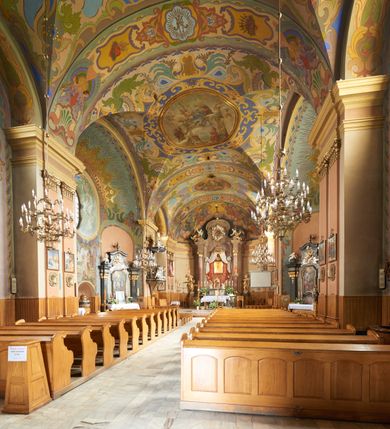 Zdjęcie nr 36: Kościół jest orientowany, murowany z cegły i kamienia, na rzucie podłużnym. Zajmuje powierzchnię około 606 metrów kwadratowych, długość bryły wynosi około 32, a szerokość 14 metrów. Jednonawowy, trójprzęsłowy korpus charakteryzuje środkowe, kwadratowe przęsło z płytkimi apsydami po bokach, wysuwającymi się przed lica elewacji bocznych, na wzór pseudotranseptu (para spłaszczonych apsyd). Przęsło wschodnie, na rzucie prostokąta, z wnękami po bokach, zakończone jest apsydą stanowiącą przestrzeń prezbiterium. Na osi kościoła i przedłużeniu prezbiterium jest piętrowa zakrystia ze skarbcem, natomiast w przedłużeniu przęsła zachodniego wieża ujęta po bokach piętrowymi lokalnościami. Korpus nawowy przekryty jest sklepieniem kolebkowym na gurtach, z lunetami, które w przęśle środkowym, na przecięciu z pseudotranseptem, tworzy rodzaj sklepienia krzyżowego z okrągłym zwierciadłem. Artykulację ścian wewnątrz kościoła stanowią pary pilastrów wspierające przerywane belkowanie. Na zewnątrz ściany korpusu, transeptu i zakrystii artykułują osadzone na niewysokim cokole lizeny wspierające obiegające całość belkowanie. Dodatkowo mury przeprute są owalnymi otworami okiennymi ujętymi profilowanymi obramieniami. Fasadę charakteryzuje w części środkowej ryzalit o skośnych ściankach bocznych oraz podziały podwójnymi i pojedynczymi pilastrami jońskimi, osadzonymi na wysokich cokołach i wspierającymi belkowanie. Do wnętrza kościoła prowadzą trzy otwory drzwiowe – w elewacji południowej, północnej i zachodniej. Drzwi w fasadzie są ujęte dekoracyjnym portalem z parą wąskich pilastrów, zwieńczonych zwężającymi się ku dołowi kapitelami, wspierających gzyms w formie łuku odcinkowego. Całość ujmuje obramienie o fantazyjnym wykroju w górnej partii, którego krawędzie obiega profilowana listwa z parą niewielkich wolut w górnych narożach. Obramienie drzwi wieńczy prosty gzyms odcinkowy nakryty blaszanym daszkiem. Po bokach wejścia znajduje się para kamiennych kropielnic. Powyżej otworu drzwiowego, pole środkowe fasady przeprute zostało oknem w kształcie stojącego prostokąta zamkniętego łukiem odcinkowym, zamurowanego w jednej trzeciej wysokości. Obramienie okna jest w formie pary pilastrów bez baz, wspierających fryz z kluczem pośrodku i parą wsporników wolutowych po bokach, dźwigających odcinkowy gzyms wieńczący. Pod parapetem umieszczono fartuch w kształcie leżącego prostokąta, o wklęsłych dolnych narożach, z dolną krawędzią zdobioną motywem lambrekinu. W skośnych ścianach bocznych ryzalitu znajduje się para zamkniętych półkoliście niszy z figurami św. Piotra po lewej i św. Pawła po prawej stronie. 
Ryzalit wieńczy trójkątny przyczółek z herbem Nowina w tympanonie. Nad belkowaniem, na całej szerokości fasady, znajduje się prosta attyka z odpowiadającymi dolnej strefie elewacji podziałami lizenowymi, wspierającymi wyłamujący się ponad nimi gzyms ozdobiony wazonami. Powyżej wznosi się górne piętro wieży, czworościenne o ściętych narożnikach, nakryte kopulastym hełmem z nasadnikiem.  
Północna, zachodnia i południowa ściana wieży przeprute są otworami okiennymi w kształcie stojącego prostokąta zamkniętego łukiem odcinkowym. Okna mają obramienia w kształcie stojących prostokątów z uszakami w górnych narożach, zwieńczone są belkowaniem w formie łuku odcinkowego z kluczem pośrodku i parą wolutowych wsporników po bokach fryzu. Podwieszenie ma formę fartucha w kształcie leżącego prostokąta o wklęsłych dolnych narożach i dolnej krawędzi zdobionej motywem lambrekinu. Powyżej otworów okiennych znajdują się okrągłe tarcze zegarowe. Pola ściętych naroży wieży zdobi ornamentyka cekinowa i kampanule. Korpus nawowy przykrywa dwuspadowy dach zakończony pięciopołaciowo nad prezbiterium i trójpołaciowo nad końcami transeptu, kryty dachówką. Na skrzyżowaniu korpusu nawowego i transeptu ulokowano obitą blachą wieżyczkę na sygnaturkę. Kolorystyka tynków kościoła utrzymana jest w odcieniu ugru, w dolnej partii różu, detal architektoniczny i ornamentyka w bieli. 

Plac przykościelny otoczony jest murowanym parkanem z usytuowanymi w nim 14 kapliczkami ze stacjami drogi krzyżowej, nakrytymi dwuspadowymi dachami. W jednym z naroży znajduje się niewielka, murowana kaplica grobowa rodziny Romerów o ceglanych ścianach artykułowanych białymi pilastrami i fasadzie ze szczytem ujętym parą spływów i zwieńczonym trójkątnym przyczółkiem.   
