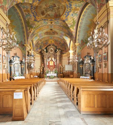 Zdjęcie nr 39: Kościół jest orientowany, murowany z cegły i kamienia, na rzucie podłużnym. Zajmuje powierzchnię około 606 metrów kwadratowych, długość bryły wynosi około 32, a szerokość 14 metrów. Jednonawowy, trójprzęsłowy korpus charakteryzuje środkowe, kwadratowe przęsło z płytkimi apsydami po bokach, wysuwającymi się przed lica elewacji bocznych, na wzór pseudotranseptu (para spłaszczonych apsyd). Przęsło wschodnie, na rzucie prostokąta, z wnękami po bokach, zakończone jest apsydą stanowiącą przestrzeń prezbiterium. Na osi kościoła i przedłużeniu prezbiterium jest piętrowa zakrystia ze skarbcem, natomiast w przedłużeniu przęsła zachodniego wieża ujęta po bokach piętrowymi lokalnościami. Korpus nawowy przekryty jest sklepieniem kolebkowym na gurtach, z lunetami, które w przęśle środkowym, na przecięciu z pseudotranseptem, tworzy rodzaj sklepienia krzyżowego z okrągłym zwierciadłem. Artykulację ścian wewnątrz kościoła stanowią pary pilastrów wspierające przerywane belkowanie. Na zewnątrz ściany korpusu, transeptu i zakrystii artykułują osadzone na niewysokim cokole lizeny wspierające obiegające całość belkowanie. Dodatkowo mury przeprute są owalnymi otworami okiennymi ujętymi profilowanymi obramieniami. Fasadę charakteryzuje w części środkowej ryzalit o skośnych ściankach bocznych oraz podziały podwójnymi i pojedynczymi pilastrami jońskimi, osadzonymi na wysokich cokołach i wspierającymi belkowanie. Do wnętrza kościoła prowadzą trzy otwory drzwiowe – w elewacji południowej, północnej i zachodniej. Drzwi w fasadzie są ujęte dekoracyjnym portalem z parą wąskich pilastrów, zwieńczonych zwężającymi się ku dołowi kapitelami, wspierających gzyms w formie łuku odcinkowego. Całość ujmuje obramienie o fantazyjnym wykroju w górnej partii, którego krawędzie obiega profilowana listwa z parą niewielkich wolut w górnych narożach. Obramienie drzwi wieńczy prosty gzyms odcinkowy nakryty blaszanym daszkiem. Po bokach wejścia znajduje się para kamiennych kropielnic. Powyżej otworu drzwiowego, pole środkowe fasady przeprute zostało oknem w kształcie stojącego prostokąta zamkniętego łukiem odcinkowym, zamurowanego w jednej trzeciej wysokości. Obramienie okna jest w formie pary pilastrów bez baz, wspierających fryz z kluczem pośrodku i parą wsporników wolutowych po bokach, dźwigających odcinkowy gzyms wieńczący. Pod parapetem umieszczono fartuch w kształcie leżącego prostokąta, o wklęsłych dolnych narożach, z dolną krawędzią zdobioną motywem lambrekinu. W skośnych ścianach bocznych ryzalitu znajduje się para zamkniętych półkoliście niszy z figurami św. Piotra po lewej i św. Pawła po prawej stronie. 
Ryzalit wieńczy trójkątny przyczółek z herbem Nowina w tympanonie. Nad belkowaniem, na całej szerokości fasady, znajduje się prosta attyka z odpowiadającymi dolnej strefie elewacji podziałami lizenowymi, wspierającymi wyłamujący się ponad nimi gzyms ozdobiony wazonami. Powyżej wznosi się górne piętro wieży, czworościenne o ściętych narożnikach, nakryte kopulastym hełmem z nasadnikiem.  
Północna, zachodnia i południowa ściana wieży przeprute są otworami okiennymi w kształcie stojącego prostokąta zamkniętego łukiem odcinkowym. Okna mają obramienia w kształcie stojących prostokątów z uszakami w górnych narożach, zwieńczone są belkowaniem w formie łuku odcinkowego z kluczem pośrodku i parą wolutowych wsporników po bokach fryzu. Podwieszenie ma formę fartucha w kształcie leżącego prostokąta o wklęsłych dolnych narożach i dolnej krawędzi zdobionej motywem lambrekinu. Powyżej otworów okiennych znajdują się okrągłe tarcze zegarowe. Pola ściętych naroży wieży zdobi ornamentyka cekinowa i kampanule. Korpus nawowy przykrywa dwuspadowy dach zakończony pięciopołaciowo nad prezbiterium i trójpołaciowo nad końcami transeptu, kryty dachówką. Na skrzyżowaniu korpusu nawowego i transeptu ulokowano obitą blachą wieżyczkę na sygnaturkę. Kolorystyka tynków kościoła utrzymana jest w odcieniu ugru, w dolnej partii różu, detal architektoniczny i ornamentyka w bieli. 

Plac przykościelny otoczony jest murowanym parkanem z usytuowanymi w nim 14 kapliczkami ze stacjami drogi krzyżowej, nakrytymi dwuspadowymi dachami. W jednym z naroży znajduje się niewielka, murowana kaplica grobowa rodziny Romerów o ceglanych ścianach artykułowanych białymi pilastrami i fasadzie ze szczytem ujętym parą spływów i zwieńczonym trójkątnym przyczółkiem.   
