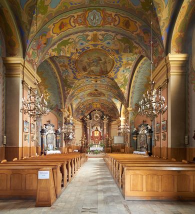 Zdjęcie nr 40: Kościół jest orientowany, murowany z cegły i kamienia, na rzucie podłużnym. Zajmuje powierzchnię około 606 metrów kwadratowych, długość bryły wynosi około 32, a szerokość 14 metrów. Jednonawowy, trójprzęsłowy korpus charakteryzuje środkowe, kwadratowe przęsło z płytkimi apsydami po bokach, wysuwającymi się przed lica elewacji bocznych, na wzór pseudotranseptu (para spłaszczonych apsyd). Przęsło wschodnie, na rzucie prostokąta, z wnękami po bokach, zakończone jest apsydą stanowiącą przestrzeń prezbiterium. Na osi kościoła i przedłużeniu prezbiterium jest piętrowa zakrystia ze skarbcem, natomiast w przedłużeniu przęsła zachodniego wieża ujęta po bokach piętrowymi lokalnościami. Korpus nawowy przekryty jest sklepieniem kolebkowym na gurtach, z lunetami, które w przęśle środkowym, na przecięciu z pseudotranseptem, tworzy rodzaj sklepienia krzyżowego z okrągłym zwierciadłem. Artykulację ścian wewnątrz kościoła stanowią pary pilastrów wspierające przerywane belkowanie. Na zewnątrz ściany korpusu, transeptu i zakrystii artykułują osadzone na niewysokim cokole lizeny wspierające obiegające całość belkowanie. Dodatkowo mury przeprute są owalnymi otworami okiennymi ujętymi profilowanymi obramieniami. Fasadę charakteryzuje w części środkowej ryzalit o skośnych ściankach bocznych oraz podziały podwójnymi i pojedynczymi pilastrami jońskimi, osadzonymi na wysokich cokołach i wspierającymi belkowanie. Do wnętrza kościoła prowadzą trzy otwory drzwiowe – w elewacji południowej, północnej i zachodniej. Drzwi w fasadzie są ujęte dekoracyjnym portalem z parą wąskich pilastrów, zwieńczonych zwężającymi się ku dołowi kapitelami, wspierających gzyms w formie łuku odcinkowego. Całość ujmuje obramienie o fantazyjnym wykroju w górnej partii, którego krawędzie obiega profilowana listwa z parą niewielkich wolut w górnych narożach. Obramienie drzwi wieńczy prosty gzyms odcinkowy nakryty blaszanym daszkiem. Po bokach wejścia znajduje się para kamiennych kropielnic. Powyżej otworu drzwiowego, pole środkowe fasady przeprute zostało oknem w kształcie stojącego prostokąta zamkniętego łukiem odcinkowym, zamurowanego w jednej trzeciej wysokości. Obramienie okna jest w formie pary pilastrów bez baz, wspierających fryz z kluczem pośrodku i parą wsporników wolutowych po bokach, dźwigających odcinkowy gzyms wieńczący. Pod parapetem umieszczono fartuch w kształcie leżącego prostokąta, o wklęsłych dolnych narożach, z dolną krawędzią zdobioną motywem lambrekinu. W skośnych ścianach bocznych ryzalitu znajduje się para zamkniętych półkoliście niszy z figurami św. Piotra po lewej i św. Pawła po prawej stronie. 
Ryzalit wieńczy trójkątny przyczółek z herbem Nowina w tympanonie. Nad belkowaniem, na całej szerokości fasady, znajduje się prosta attyka z odpowiadającymi dolnej strefie elewacji podziałami lizenowymi, wspierającymi wyłamujący się ponad nimi gzyms ozdobiony wazonami. Powyżej wznosi się górne piętro wieży, czworościenne o ściętych narożnikach, nakryte kopulastym hełmem z nasadnikiem.  
Północna, zachodnia i południowa ściana wieży przeprute są otworami okiennymi w kształcie stojącego prostokąta zamkniętego łukiem odcinkowym. Okna mają obramienia w kształcie stojących prostokątów z uszakami w górnych narożach, zwieńczone są belkowaniem w formie łuku odcinkowego z kluczem pośrodku i parą wolutowych wsporników po bokach fryzu. Podwieszenie ma formę fartucha w kształcie leżącego prostokąta o wklęsłych dolnych narożach i dolnej krawędzi zdobionej motywem lambrekinu. Powyżej otworów okiennych znajdują się okrągłe tarcze zegarowe. Pola ściętych naroży wieży zdobi ornamentyka cekinowa i kampanule. Korpus nawowy przykrywa dwuspadowy dach zakończony pięciopołaciowo nad prezbiterium i trójpołaciowo nad końcami transeptu, kryty dachówką. Na skrzyżowaniu korpusu nawowego i transeptu ulokowano obitą blachą wieżyczkę na sygnaturkę. Kolorystyka tynków kościoła utrzymana jest w odcieniu ugru, w dolnej partii różu, detal architektoniczny i ornamentyka w bieli. 

Plac przykościelny otoczony jest murowanym parkanem z usytuowanymi w nim 14 kapliczkami ze stacjami drogi krzyżowej, nakrytymi dwuspadowymi dachami. W jednym z naroży znajduje się niewielka, murowana kaplica grobowa rodziny Romerów o ceglanych ścianach artykułowanych białymi pilastrami i fasadzie ze szczytem ujętym parą spływów i zwieńczonym trójkątnym przyczółkiem.   
