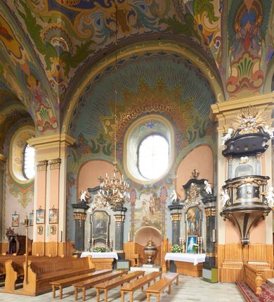 Zdjęcie nr 8: Kościół jest orientowany, murowany z cegły i kamienia, na rzucie podłużnym. Zajmuje powierzchnię około 606 metrów kwadratowych, długość bryły wynosi około 32, a szerokość 14 metrów. Jednonawowy, trójprzęsłowy korpus charakteryzuje środkowe, kwadratowe przęsło z płytkimi apsydami po bokach, wysuwającymi się przed lica elewacji bocznych, na wzór pseudotranseptu (para spłaszczonych apsyd). Przęsło wschodnie, na rzucie prostokąta, z wnękami po bokach, zakończone jest apsydą stanowiącą przestrzeń prezbiterium. Na osi kościoła i przedłużeniu prezbiterium jest piętrowa zakrystia ze skarbcem, natomiast w przedłużeniu przęsła zachodniego wieża ujęta po bokach piętrowymi lokalnościami. Korpus nawowy przekryty jest sklepieniem kolebkowym na gurtach, z lunetami, które w przęśle środkowym, na przecięciu z pseudotranseptem, tworzy rodzaj sklepienia krzyżowego z okrągłym zwierciadłem. Artykulację ścian wewnątrz kościoła stanowią pary pilastrów wspierające przerywane belkowanie. Na zewnątrz ściany korpusu, transeptu i zakrystii artykułują osadzone na niewysokim cokole lizeny wspierające obiegające całość belkowanie. Dodatkowo mury przeprute są owalnymi otworami okiennymi ujętymi profilowanymi obramieniami. Fasadę charakteryzuje w części środkowej ryzalit o skośnych ściankach bocznych oraz podziały podwójnymi i pojedynczymi pilastrami jońskimi, osadzonymi na wysokich cokołach i wspierającymi belkowanie. Do wnętrza kościoła prowadzą trzy otwory drzwiowe – w elewacji południowej, północnej i zachodniej. Drzwi w fasadzie są ujęte dekoracyjnym portalem z parą wąskich pilastrów, zwieńczonych zwężającymi się ku dołowi kapitelami, wspierających gzyms w formie łuku odcinkowego. Całość ujmuje obramienie o fantazyjnym wykroju w górnej partii, którego krawędzie obiega profilowana listwa z parą niewielkich wolut w górnych narożach. Obramienie drzwi wieńczy prosty gzyms odcinkowy nakryty blaszanym daszkiem. Po bokach wejścia znajduje się para kamiennych kropielnic. Powyżej otworu drzwiowego, pole środkowe fasady przeprute zostało oknem w kształcie stojącego prostokąta zamkniętego łukiem odcinkowym, zamurowanego w jednej trzeciej wysokości. Obramienie okna jest w formie pary pilastrów bez baz, wspierających fryz z kluczem pośrodku i parą wsporników wolutowych po bokach, dźwigających odcinkowy gzyms wieńczący. Pod parapetem umieszczono fartuch w kształcie leżącego prostokąta, o wklęsłych dolnych narożach, z dolną krawędzią zdobioną motywem lambrekinu. W skośnych ścianach bocznych ryzalitu znajduje się para zamkniętych półkoliście niszy z figurami św. Piotra po lewej i św. Pawła po prawej stronie. 
Ryzalit wieńczy trójkątny przyczółek z herbem Nowina w tympanonie. Nad belkowaniem, na całej szerokości fasady, znajduje się prosta attyka z odpowiadającymi dolnej strefie elewacji podziałami lizenowymi, wspierającymi wyłamujący się ponad nimi gzyms ozdobiony wazonami. Powyżej wznosi się górne piętro wieży, czworościenne o ściętych narożnikach, nakryte kopulastym hełmem z nasadnikiem.  
Północna, zachodnia i południowa ściana wieży przeprute są otworami okiennymi w kształcie stojącego prostokąta zamkniętego łukiem odcinkowym. Okna mają obramienia w kształcie stojących prostokątów z uszakami w górnych narożach, zwieńczone są belkowaniem w formie łuku odcinkowego z kluczem pośrodku i parą wolutowych wsporników po bokach fryzu. Podwieszenie ma formę fartucha w kształcie leżącego prostokąta o wklęsłych dolnych narożach i dolnej krawędzi zdobionej motywem lambrekinu. Powyżej otworów okiennych znajdują się okrągłe tarcze zegarowe. Pola ściętych naroży wieży zdobi ornamentyka cekinowa i kampanule. Korpus nawowy przykrywa dwuspadowy dach zakończony pięciopołaciowo nad prezbiterium i trójpołaciowo nad końcami transeptu, kryty dachówką. Na skrzyżowaniu korpusu nawowego i transeptu ulokowano obitą blachą wieżyczkę na sygnaturkę. Kolorystyka tynków kościoła utrzymana jest w odcieniu ugru, w dolnej partii różu, detal architektoniczny i ornamentyka w bieli. 

Plac przykościelny otoczony jest murowanym parkanem z usytuowanymi w nim 14 kapliczkami ze stacjami drogi krzyżowej, nakrytymi dwuspadowymi dachami. W jednym z naroży znajduje się niewielka, murowana kaplica grobowa rodziny Romerów o ceglanych ścianach artykułowanych białymi pilastrami i fasadzie ze szczytem ujętym parą spływów i zwieńczonym trójkątnym przyczółkiem.   
