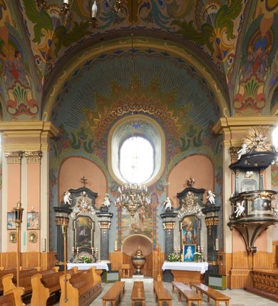 Zdjęcie nr 9: Kościół jest orientowany, murowany z cegły i kamienia, na rzucie podłużnym. Zajmuje powierzchnię około 606 metrów kwadratowych, długość bryły wynosi około 32, a szerokość 14 metrów. Jednonawowy, trójprzęsłowy korpus charakteryzuje środkowe, kwadratowe przęsło z płytkimi apsydami po bokach, wysuwającymi się przed lica elewacji bocznych, na wzór pseudotranseptu (para spłaszczonych apsyd). Przęsło wschodnie, na rzucie prostokąta, z wnękami po bokach, zakończone jest apsydą stanowiącą przestrzeń prezbiterium. Na osi kościoła i przedłużeniu prezbiterium jest piętrowa zakrystia ze skarbcem, natomiast w przedłużeniu przęsła zachodniego wieża ujęta po bokach piętrowymi lokalnościami. Korpus nawowy przekryty jest sklepieniem kolebkowym na gurtach, z lunetami, które w przęśle środkowym, na przecięciu z pseudotranseptem, tworzy rodzaj sklepienia krzyżowego z okrągłym zwierciadłem. Artykulację ścian wewnątrz kościoła stanowią pary pilastrów wspierające przerywane belkowanie. Na zewnątrz ściany korpusu, transeptu i zakrystii artykułują osadzone na niewysokim cokole lizeny wspierające obiegające całość belkowanie. Dodatkowo mury przeprute są owalnymi otworami okiennymi ujętymi profilowanymi obramieniami. Fasadę charakteryzuje w części środkowej ryzalit o skośnych ściankach bocznych oraz podziały podwójnymi i pojedynczymi pilastrami jońskimi, osadzonymi na wysokich cokołach i wspierającymi belkowanie. Do wnętrza kościoła prowadzą trzy otwory drzwiowe – w elewacji południowej, północnej i zachodniej. Drzwi w fasadzie są ujęte dekoracyjnym portalem z parą wąskich pilastrów, zwieńczonych zwężającymi się ku dołowi kapitelami, wspierających gzyms w formie łuku odcinkowego. Całość ujmuje obramienie o fantazyjnym wykroju w górnej partii, którego krawędzie obiega profilowana listwa z parą niewielkich wolut w górnych narożach. Obramienie drzwi wieńczy prosty gzyms odcinkowy nakryty blaszanym daszkiem. Po bokach wejścia znajduje się para kamiennych kropielnic. Powyżej otworu drzwiowego, pole środkowe fasady przeprute zostało oknem w kształcie stojącego prostokąta zamkniętego łukiem odcinkowym, zamurowanego w jednej trzeciej wysokości. Obramienie okna jest w formie pary pilastrów bez baz, wspierających fryz z kluczem pośrodku i parą wsporników wolutowych po bokach, dźwigających odcinkowy gzyms wieńczący. Pod parapetem umieszczono fartuch w kształcie leżącego prostokąta, o wklęsłych dolnych narożach, z dolną krawędzią zdobioną motywem lambrekinu. W skośnych ścianach bocznych ryzalitu znajduje się para zamkniętych półkoliście niszy z figurami św. Piotra po lewej i św. Pawła po prawej stronie. 
Ryzalit wieńczy trójkątny przyczółek z herbem Nowina w tympanonie. Nad belkowaniem, na całej szerokości fasady, znajduje się prosta attyka z odpowiadającymi dolnej strefie elewacji podziałami lizenowymi, wspierającymi wyłamujący się ponad nimi gzyms ozdobiony wazonami. Powyżej wznosi się górne piętro wieży, czworościenne o ściętych narożnikach, nakryte kopulastym hełmem z nasadnikiem.  
Północna, zachodnia i południowa ściana wieży przeprute są otworami okiennymi w kształcie stojącego prostokąta zamkniętego łukiem odcinkowym. Okna mają obramienia w kształcie stojących prostokątów z uszakami w górnych narożach, zwieńczone są belkowaniem w formie łuku odcinkowego z kluczem pośrodku i parą wolutowych wsporników po bokach fryzu. Podwieszenie ma formę fartucha w kształcie leżącego prostokąta o wklęsłych dolnych narożach i dolnej krawędzi zdobionej motywem lambrekinu. Powyżej otworów okiennych znajdują się okrągłe tarcze zegarowe. Pola ściętych naroży wieży zdobi ornamentyka cekinowa i kampanule. Korpus nawowy przykrywa dwuspadowy dach zakończony pięciopołaciowo nad prezbiterium i trójpołaciowo nad końcami transeptu, kryty dachówką. Na skrzyżowaniu korpusu nawowego i transeptu ulokowano obitą blachą wieżyczkę na sygnaturkę. Kolorystyka tynków kościoła utrzymana jest w odcieniu ugru, w dolnej partii różu, detal architektoniczny i ornamentyka w bieli. 

Plac przykościelny otoczony jest murowanym parkanem z usytuowanymi w nim 14 kapliczkami ze stacjami drogi krzyżowej, nakrytymi dwuspadowymi dachami. W jednym z naroży znajduje się niewielka, murowana kaplica grobowa rodziny Romerów o ceglanych ścianach artykułowanych białymi pilastrami i fasadzie ze szczytem ujętym parą spływów i zwieńczonym trójkątnym przyczółkiem.   
