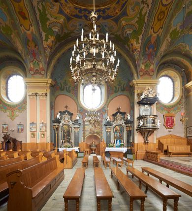 Zdjęcie nr 11: Kościół jest orientowany, murowany z cegły i kamienia, na rzucie podłużnym. Zajmuje powierzchnię około 606 metrów kwadratowych, długość bryły wynosi około 32, a szerokość 14 metrów. Jednonawowy, trójprzęsłowy korpus charakteryzuje środkowe, kwadratowe przęsło z płytkimi apsydami po bokach, wysuwającymi się przed lica elewacji bocznych, na wzór pseudotranseptu (para spłaszczonych apsyd). Przęsło wschodnie, na rzucie prostokąta, z wnękami po bokach, zakończone jest apsydą stanowiącą przestrzeń prezbiterium. Na osi kościoła i przedłużeniu prezbiterium jest piętrowa zakrystia ze skarbcem, natomiast w przedłużeniu przęsła zachodniego wieża ujęta po bokach piętrowymi lokalnościami. Korpus nawowy przekryty jest sklepieniem kolebkowym na gurtach, z lunetami, które w przęśle środkowym, na przecięciu z pseudotranseptem, tworzy rodzaj sklepienia krzyżowego z okrągłym zwierciadłem. Artykulację ścian wewnątrz kościoła stanowią pary pilastrów wspierające przerywane belkowanie. Na zewnątrz ściany korpusu, transeptu i zakrystii artykułują osadzone na niewysokim cokole lizeny wspierające obiegające całość belkowanie. Dodatkowo mury przeprute są owalnymi otworami okiennymi ujętymi profilowanymi obramieniami. Fasadę charakteryzuje w części środkowej ryzalit o skośnych ściankach bocznych oraz podziały podwójnymi i pojedynczymi pilastrami jońskimi, osadzonymi na wysokich cokołach i wspierającymi belkowanie. Do wnętrza kościoła prowadzą trzy otwory drzwiowe – w elewacji południowej, północnej i zachodniej. Drzwi w fasadzie są ujęte dekoracyjnym portalem z parą wąskich pilastrów, zwieńczonych zwężającymi się ku dołowi kapitelami, wspierających gzyms w formie łuku odcinkowego. Całość ujmuje obramienie o fantazyjnym wykroju w górnej partii, którego krawędzie obiega profilowana listwa z parą niewielkich wolut w górnych narożach. Obramienie drzwi wieńczy prosty gzyms odcinkowy nakryty blaszanym daszkiem. Po bokach wejścia znajduje się para kamiennych kropielnic. Powyżej otworu drzwiowego, pole środkowe fasady przeprute zostało oknem w kształcie stojącego prostokąta zamkniętego łukiem odcinkowym, zamurowanego w jednej trzeciej wysokości. Obramienie okna jest w formie pary pilastrów bez baz, wspierających fryz z kluczem pośrodku i parą wsporników wolutowych po bokach, dźwigających odcinkowy gzyms wieńczący. Pod parapetem umieszczono fartuch w kształcie leżącego prostokąta, o wklęsłych dolnych narożach, z dolną krawędzią zdobioną motywem lambrekinu. W skośnych ścianach bocznych ryzalitu znajduje się para zamkniętych półkoliście niszy z figurami św. Piotra po lewej i św. Pawła po prawej stronie. 
Ryzalit wieńczy trójkątny przyczółek z herbem Nowina w tympanonie. Nad belkowaniem, na całej szerokości fasady, znajduje się prosta attyka z odpowiadającymi dolnej strefie elewacji podziałami lizenowymi, wspierającymi wyłamujący się ponad nimi gzyms ozdobiony wazonami. Powyżej wznosi się górne piętro wieży, czworościenne o ściętych narożnikach, nakryte kopulastym hełmem z nasadnikiem.  
Północna, zachodnia i południowa ściana wieży przeprute są otworami okiennymi w kształcie stojącego prostokąta zamkniętego łukiem odcinkowym. Okna mają obramienia w kształcie stojących prostokątów z uszakami w górnych narożach, zwieńczone są belkowaniem w formie łuku odcinkowego z kluczem pośrodku i parą wolutowych wsporników po bokach fryzu. Podwieszenie ma formę fartucha w kształcie leżącego prostokąta o wklęsłych dolnych narożach i dolnej krawędzi zdobionej motywem lambrekinu. Powyżej otworów okiennych znajdują się okrągłe tarcze zegarowe. Pola ściętych naroży wieży zdobi ornamentyka cekinowa i kampanule. Korpus nawowy przykrywa dwuspadowy dach zakończony pięciopołaciowo nad prezbiterium i trójpołaciowo nad końcami transeptu, kryty dachówką. Na skrzyżowaniu korpusu nawowego i transeptu ulokowano obitą blachą wieżyczkę na sygnaturkę. Kolorystyka tynków kościoła utrzymana jest w odcieniu ugru, w dolnej partii różu, detal architektoniczny i ornamentyka w bieli. 

Plac przykościelny otoczony jest murowanym parkanem z usytuowanymi w nim 14 kapliczkami ze stacjami drogi krzyżowej, nakrytymi dwuspadowymi dachami. W jednym z naroży znajduje się niewielka, murowana kaplica grobowa rodziny Romerów o ceglanych ścianach artykułowanych białymi pilastrami i fasadzie ze szczytem ujętym parą spływów i zwieńczonym trójkątnym przyczółkiem.   
