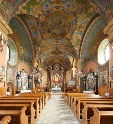 Zdjęcie nr 34: Kościół jest orientowany, murowany z cegły i kamienia, na rzucie podłużnym. Zajmuje powierzchnię około 606 metrów kwadratowych, długość bryły wynosi około 32, a szerokość 14 metrów. Jednonawowy, trójprzęsłowy korpus charakteryzuje środkowe, kwadratowe przęsło z płytkimi apsydami po bokach, wysuwającymi się przed lica elewacji bocznych, na wzór pseudotranseptu (para spłaszczonych apsyd). Przęsło wschodnie, na rzucie prostokąta, z wnękami po bokach, zakończone jest apsydą stanowiącą przestrzeń prezbiterium. Na osi kościoła i przedłużeniu prezbiterium jest piętrowa zakrystia ze skarbcem, natomiast w przedłużeniu przęsła zachodniego wieża ujęta po bokach piętrowymi lokalnościami. Korpus nawowy przekryty jest sklepieniem kolebkowym na gurtach, z lunetami, które w przęśle środkowym, na przecięciu z pseudotranseptem, tworzy rodzaj sklepienia krzyżowego z okrągłym zwierciadłem. Artykulację ścian wewnątrz kościoła stanowią pary pilastrów wspierające przerywane belkowanie. Na zewnątrz ściany korpusu, transeptu i zakrystii artykułują osadzone na niewysokim cokole lizeny wspierające obiegające całość belkowanie. Dodatkowo mury przeprute są owalnymi otworami okiennymi ujętymi profilowanymi obramieniami. Fasadę charakteryzuje w części środkowej ryzalit o skośnych ściankach bocznych oraz podziały podwójnymi i pojedynczymi pilastrami jońskimi, osadzonymi na wysokich cokołach i wspierającymi belkowanie. Do wnętrza kościoła prowadzą trzy otwory drzwiowe – w elewacji południowej, północnej i zachodniej. Drzwi w fasadzie są ujęte dekoracyjnym portalem z parą wąskich pilastrów, zwieńczonych zwężającymi się ku dołowi kapitelami, wspierających gzyms w formie łuku odcinkowego. Całość ujmuje obramienie o fantazyjnym wykroju w górnej partii, którego krawędzie obiega profilowana listwa z parą niewielkich wolut w górnych narożach. Obramienie drzwi wieńczy prosty gzyms odcinkowy nakryty blaszanym daszkiem. Po bokach wejścia znajduje się para kamiennych kropielnic. Powyżej otworu drzwiowego, pole środkowe fasady przeprute zostało oknem w kształcie stojącego prostokąta zamkniętego łukiem odcinkowym, zamurowanego w jednej trzeciej wysokości. Obramienie okna jest w formie pary pilastrów bez baz, wspierających fryz z kluczem pośrodku i parą wsporników wolutowych po bokach, dźwigających odcinkowy gzyms wieńczący. Pod parapetem umieszczono fartuch w kształcie leżącego prostokąta, o wklęsłych dolnych narożach, z dolną krawędzią zdobioną motywem lambrekinu. W skośnych ścianach bocznych ryzalitu znajduje się para zamkniętych półkoliście niszy z figurami św. Piotra po lewej i św. Pawła po prawej stronie. 
Ryzalit wieńczy trójkątny przyczółek z herbem Nowina w tympanonie. Nad belkowaniem, na całej szerokości fasady, znajduje się prosta attyka z odpowiadającymi dolnej strefie elewacji podziałami lizenowymi, wspierającymi wyłamujący się ponad nimi gzyms ozdobiony wazonami. Powyżej wznosi się górne piętro wieży, czworościenne o ściętych narożnikach, nakryte kopulastym hełmem z nasadnikiem.  
Północna, zachodnia i południowa ściana wieży przeprute są otworami okiennymi w kształcie stojącego prostokąta zamkniętego łukiem odcinkowym. Okna mają obramienia w kształcie stojących prostokątów z uszakami w górnych narożach, zwieńczone są belkowaniem w formie łuku odcinkowego z kluczem pośrodku i parą wolutowych wsporników po bokach fryzu. Podwieszenie ma formę fartucha w kształcie leżącego prostokąta o wklęsłych dolnych narożach i dolnej krawędzi zdobionej motywem lambrekinu. Powyżej otworów okiennych znajdują się okrągłe tarcze zegarowe. Pola ściętych naroży wieży zdobi ornamentyka cekinowa i kampanule. Korpus nawowy przykrywa dwuspadowy dach zakończony pięciopołaciowo nad prezbiterium i trójpołaciowo nad końcami transeptu, kryty dachówką. Na skrzyżowaniu korpusu nawowego i transeptu ulokowano obitą blachą wieżyczkę na sygnaturkę. Kolorystyka tynków kościoła utrzymana jest w odcieniu ugru, w dolnej partii różu, detal architektoniczny i ornamentyka w bieli. 

Plac przykościelny otoczony jest murowanym parkanem z usytuowanymi w nim 14 kapliczkami ze stacjami drogi krzyżowej, nakrytymi dwuspadowymi dachami. W jednym z naroży znajduje się niewielka, murowana kaplica grobowa rodziny Romerów o ceglanych ścianach artykułowanych białymi pilastrami i fasadzie ze szczytem ujętym parą spływów i zwieńczonym trójkątnym przyczółkiem.   
