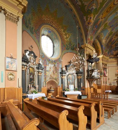 Zdjęcie nr 12: Kościół jest orientowany, murowany z cegły i kamienia, na rzucie podłużnym. Zajmuje powierzchnię około 606 metrów kwadratowych, długość bryły wynosi około 32, a szerokość 14 metrów. Jednonawowy, trójprzęsłowy korpus charakteryzuje środkowe, kwadratowe przęsło z płytkimi apsydami po bokach, wysuwającymi się przed lica elewacji bocznych, na wzór pseudotranseptu (para spłaszczonych apsyd). Przęsło wschodnie, na rzucie prostokąta, z wnękami po bokach, zakończone jest apsydą stanowiącą przestrzeń prezbiterium. Na osi kościoła i przedłużeniu prezbiterium jest piętrowa zakrystia ze skarbcem, natomiast w przedłużeniu przęsła zachodniego wieża ujęta po bokach piętrowymi lokalnościami. Korpus nawowy przekryty jest sklepieniem kolebkowym na gurtach, z lunetami, które w przęśle środkowym, na przecięciu z pseudotranseptem, tworzy rodzaj sklepienia krzyżowego z okrągłym zwierciadłem. Artykulację ścian wewnątrz kościoła stanowią pary pilastrów wspierające przerywane belkowanie. Na zewnątrz ściany korpusu, transeptu i zakrystii artykułują osadzone na niewysokim cokole lizeny wspierające obiegające całość belkowanie. Dodatkowo mury przeprute są owalnymi otworami okiennymi ujętymi profilowanymi obramieniami. Fasadę charakteryzuje w części środkowej ryzalit o skośnych ściankach bocznych oraz podziały podwójnymi i pojedynczymi pilastrami jońskimi, osadzonymi na wysokich cokołach i wspierającymi belkowanie. Do wnętrza kościoła prowadzą trzy otwory drzwiowe – w elewacji południowej, północnej i zachodniej. Drzwi w fasadzie są ujęte dekoracyjnym portalem z parą wąskich pilastrów, zwieńczonych zwężającymi się ku dołowi kapitelami, wspierających gzyms w formie łuku odcinkowego. Całość ujmuje obramienie o fantazyjnym wykroju w górnej partii, którego krawędzie obiega profilowana listwa z parą niewielkich wolut w górnych narożach. Obramienie drzwi wieńczy prosty gzyms odcinkowy nakryty blaszanym daszkiem. Po bokach wejścia znajduje się para kamiennych kropielnic. Powyżej otworu drzwiowego, pole środkowe fasady przeprute zostało oknem w kształcie stojącego prostokąta zamkniętego łukiem odcinkowym, zamurowanego w jednej trzeciej wysokości. Obramienie okna jest w formie pary pilastrów bez baz, wspierających fryz z kluczem pośrodku i parą wsporników wolutowych po bokach, dźwigających odcinkowy gzyms wieńczący. Pod parapetem umieszczono fartuch w kształcie leżącego prostokąta, o wklęsłych dolnych narożach, z dolną krawędzią zdobioną motywem lambrekinu. W skośnych ścianach bocznych ryzalitu znajduje się para zamkniętych półkoliście niszy z figurami św. Piotra po lewej i św. Pawła po prawej stronie. 
Ryzalit wieńczy trójkątny przyczółek z herbem Nowina w tympanonie. Nad belkowaniem, na całej szerokości fasady, znajduje się prosta attyka z odpowiadającymi dolnej strefie elewacji podziałami lizenowymi, wspierającymi wyłamujący się ponad nimi gzyms ozdobiony wazonami. Powyżej wznosi się górne piętro wieży, czworościenne o ściętych narożnikach, nakryte kopulastym hełmem z nasadnikiem.  
Północna, zachodnia i południowa ściana wieży przeprute są otworami okiennymi w kształcie stojącego prostokąta zamkniętego łukiem odcinkowym. Okna mają obramienia w kształcie stojących prostokątów z uszakami w górnych narożach, zwieńczone są belkowaniem w formie łuku odcinkowego z kluczem pośrodku i parą wolutowych wsporników po bokach fryzu. Podwieszenie ma formę fartucha w kształcie leżącego prostokąta o wklęsłych dolnych narożach i dolnej krawędzi zdobionej motywem lambrekinu. Powyżej otworów okiennych znajdują się okrągłe tarcze zegarowe. Pola ściętych naroży wieży zdobi ornamentyka cekinowa i kampanule. Korpus nawowy przykrywa dwuspadowy dach zakończony pięciopołaciowo nad prezbiterium i trójpołaciowo nad końcami transeptu, kryty dachówką. Na skrzyżowaniu korpusu nawowego i transeptu ulokowano obitą blachą wieżyczkę na sygnaturkę. Kolorystyka tynków kościoła utrzymana jest w odcieniu ugru, w dolnej partii różu, detal architektoniczny i ornamentyka w bieli. 

Plac przykościelny otoczony jest murowanym parkanem z usytuowanymi w nim 14 kapliczkami ze stacjami drogi krzyżowej, nakrytymi dwuspadowymi dachami. W jednym z naroży znajduje się niewielka, murowana kaplica grobowa rodziny Romerów o ceglanych ścianach artykułowanych białymi pilastrami i fasadzie ze szczytem ujętym parą spływów i zwieńczonym trójkątnym przyczółkiem.   
