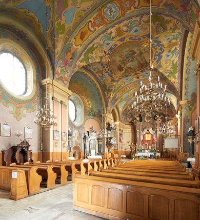 Zdjęcie nr 5: Kościół jest orientowany, murowany z cegły i kamienia, na rzucie podłużnym. Zajmuje powierzchnię około 606 metrów kwadratowych, długość bryły wynosi około 32, a szerokość 14 metrów. Jednonawowy, trójprzęsłowy korpus charakteryzuje środkowe, kwadratowe przęsło z płytkimi apsydami po bokach, wysuwającymi się przed lica elewacji bocznych, na wzór pseudotranseptu (para spłaszczonych apsyd). Przęsło wschodnie, na rzucie prostokąta, z wnękami po bokach, zakończone jest apsydą stanowiącą przestrzeń prezbiterium. Na osi kościoła i przedłużeniu prezbiterium jest piętrowa zakrystia ze skarbcem, natomiast w przedłużeniu przęsła zachodniego wieża ujęta po bokach piętrowymi lokalnościami. Korpus nawowy przekryty jest sklepieniem kolebkowym na gurtach, z lunetami, które w przęśle środkowym, na przecięciu z pseudotranseptem, tworzy rodzaj sklepienia krzyżowego z okrągłym zwierciadłem. Artykulację ścian wewnątrz kościoła stanowią pary pilastrów wspierające przerywane belkowanie. Na zewnątrz ściany korpusu, transeptu i zakrystii artykułują osadzone na niewysokim cokole lizeny wspierające obiegające całość belkowanie. Dodatkowo mury przeprute są owalnymi otworami okiennymi ujętymi profilowanymi obramieniami. Fasadę charakteryzuje w części środkowej ryzalit o skośnych ściankach bocznych oraz podziały podwójnymi i pojedynczymi pilastrami jońskimi, osadzonymi na wysokich cokołach i wspierającymi belkowanie. Do wnętrza kościoła prowadzą trzy otwory drzwiowe – w elewacji południowej, północnej i zachodniej. Drzwi w fasadzie są ujęte dekoracyjnym portalem z parą wąskich pilastrów, zwieńczonych zwężającymi się ku dołowi kapitelami, wspierających gzyms w formie łuku odcinkowego. Całość ujmuje obramienie o fantazyjnym wykroju w górnej partii, którego krawędzie obiega profilowana listwa z parą niewielkich wolut w górnych narożach. Obramienie drzwi wieńczy prosty gzyms odcinkowy nakryty blaszanym daszkiem. Po bokach wejścia znajduje się para kamiennych kropielnic. Powyżej otworu drzwiowego, pole środkowe fasady przeprute zostało oknem w kształcie stojącego prostokąta zamkniętego łukiem odcinkowym, zamurowanego w jednej trzeciej wysokości. Obramienie okna jest w formie pary pilastrów bez baz, wspierających fryz z kluczem pośrodku i parą wsporników wolutowych po bokach, dźwigających odcinkowy gzyms wieńczący. Pod parapetem umieszczono fartuch w kształcie leżącego prostokąta, o wklęsłych dolnych narożach, z dolną krawędzią zdobioną motywem lambrekinu. W skośnych ścianach bocznych ryzalitu znajduje się para zamkniętych półkoliście niszy z figurami św. Piotra po lewej i św. Pawła po prawej stronie. 
Ryzalit wieńczy trójkątny przyczółek z herbem Nowina w tympanonie. Nad belkowaniem, na całej szerokości fasady, znajduje się prosta attyka z odpowiadającymi dolnej strefie elewacji podziałami lizenowymi, wspierającymi wyłamujący się ponad nimi gzyms ozdobiony wazonami. Powyżej wznosi się górne piętro wieży, czworościenne o ściętych narożnikach, nakryte kopulastym hełmem z nasadnikiem.  
Północna, zachodnia i południowa ściana wieży przeprute są otworami okiennymi w kształcie stojącego prostokąta zamkniętego łukiem odcinkowym. Okna mają obramienia w kształcie stojących prostokątów z uszakami w górnych narożach, zwieńczone są belkowaniem w formie łuku odcinkowego z kluczem pośrodku i parą wolutowych wsporników po bokach fryzu. Podwieszenie ma formę fartucha w kształcie leżącego prostokąta o wklęsłych dolnych narożach i dolnej krawędzi zdobionej motywem lambrekinu. Powyżej otworów okiennych znajdują się okrągłe tarcze zegarowe. Pola ściętych naroży wieży zdobi ornamentyka cekinowa i kampanule. Korpus nawowy przykrywa dwuspadowy dach zakończony pięciopołaciowo nad prezbiterium i trójpołaciowo nad końcami transeptu, kryty dachówką. Na skrzyżowaniu korpusu nawowego i transeptu ulokowano obitą blachą wieżyczkę na sygnaturkę. Kolorystyka tynków kościoła utrzymana jest w odcieniu ugru, w dolnej partii różu, detal architektoniczny i ornamentyka w bieli. 

Plac przykościelny otoczony jest murowanym parkanem z usytuowanymi w nim 14 kapliczkami ze stacjami drogi krzyżowej, nakrytymi dwuspadowymi dachami. W jednym z naroży znajduje się niewielka, murowana kaplica grobowa rodziny Romerów o ceglanych ścianach artykułowanych białymi pilastrami i fasadzie ze szczytem ujętym parą spływów i zwieńczonym trójkątnym przyczółkiem.   
