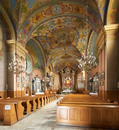 Zdjęcie nr 42: Kościół jest orientowany, murowany z cegły i kamienia, na rzucie podłużnym. Zajmuje powierzchnię około 606 metrów kwadratowych, długość bryły wynosi około 32, a szerokość 14 metrów. Jednonawowy, trójprzęsłowy korpus charakteryzuje środkowe, kwadratowe przęsło z płytkimi apsydami po bokach, wysuwającymi się przed lica elewacji bocznych, na wzór pseudotranseptu (para spłaszczonych apsyd). Przęsło wschodnie, na rzucie prostokąta, z wnękami po bokach, zakończone jest apsydą stanowiącą przestrzeń prezbiterium. Na osi kościoła i przedłużeniu prezbiterium jest piętrowa zakrystia ze skarbcem, natomiast w przedłużeniu przęsła zachodniego wieża ujęta po bokach piętrowymi lokalnościami. Korpus nawowy przekryty jest sklepieniem kolebkowym na gurtach, z lunetami, które w przęśle środkowym, na przecięciu z pseudotranseptem, tworzy rodzaj sklepienia krzyżowego z okrągłym zwierciadłem. Artykulację ścian wewnątrz kościoła stanowią pary pilastrów wspierające przerywane belkowanie. Na zewnątrz ściany korpusu, transeptu i zakrystii artykułują osadzone na niewysokim cokole lizeny wspierające obiegające całość belkowanie. Dodatkowo mury przeprute są owalnymi otworami okiennymi ujętymi profilowanymi obramieniami. Fasadę charakteryzuje w części środkowej ryzalit o skośnych ściankach bocznych oraz podziały podwójnymi i pojedynczymi pilastrami jońskimi, osadzonymi na wysokich cokołach i wspierającymi belkowanie. Do wnętrza kościoła prowadzą trzy otwory drzwiowe – w elewacji południowej, północnej i zachodniej. Drzwi w fasadzie są ujęte dekoracyjnym portalem z parą wąskich pilastrów, zwieńczonych zwężającymi się ku dołowi kapitelami, wspierających gzyms w formie łuku odcinkowego. Całość ujmuje obramienie o fantazyjnym wykroju w górnej partii, którego krawędzie obiega profilowana listwa z parą niewielkich wolut w górnych narożach. Obramienie drzwi wieńczy prosty gzyms odcinkowy nakryty blaszanym daszkiem. Po bokach wejścia znajduje się para kamiennych kropielnic. Powyżej otworu drzwiowego, pole środkowe fasady przeprute zostało oknem w kształcie stojącego prostokąta zamkniętego łukiem odcinkowym, zamurowanego w jednej trzeciej wysokości. Obramienie okna jest w formie pary pilastrów bez baz, wspierających fryz z kluczem pośrodku i parą wsporników wolutowych po bokach, dźwigających odcinkowy gzyms wieńczący. Pod parapetem umieszczono fartuch w kształcie leżącego prostokąta, o wklęsłych dolnych narożach, z dolną krawędzią zdobioną motywem lambrekinu. W skośnych ścianach bocznych ryzalitu znajduje się para zamkniętych półkoliście niszy z figurami św. Piotra po lewej i św. Pawła po prawej stronie. 
Ryzalit wieńczy trójkątny przyczółek z herbem Nowina w tympanonie. Nad belkowaniem, na całej szerokości fasady, znajduje się prosta attyka z odpowiadającymi dolnej strefie elewacji podziałami lizenowymi, wspierającymi wyłamujący się ponad nimi gzyms ozdobiony wazonami. Powyżej wznosi się górne piętro wieży, czworościenne o ściętych narożnikach, nakryte kopulastym hełmem z nasadnikiem.  
Północna, zachodnia i południowa ściana wieży przeprute są otworami okiennymi w kształcie stojącego prostokąta zamkniętego łukiem odcinkowym. Okna mają obramienia w kształcie stojących prostokątów z uszakami w górnych narożach, zwieńczone są belkowaniem w formie łuku odcinkowego z kluczem pośrodku i parą wolutowych wsporników po bokach fryzu. Podwieszenie ma formę fartucha w kształcie leżącego prostokąta o wklęsłych dolnych narożach i dolnej krawędzi zdobionej motywem lambrekinu. Powyżej otworów okiennych znajdują się okrągłe tarcze zegarowe. Pola ściętych naroży wieży zdobi ornamentyka cekinowa i kampanule. Korpus nawowy przykrywa dwuspadowy dach zakończony pięciopołaciowo nad prezbiterium i trójpołaciowo nad końcami transeptu, kryty dachówką. Na skrzyżowaniu korpusu nawowego i transeptu ulokowano obitą blachą wieżyczkę na sygnaturkę. Kolorystyka tynków kościoła utrzymana jest w odcieniu ugru, w dolnej partii różu, detal architektoniczny i ornamentyka w bieli. 

Plac przykościelny otoczony jest murowanym parkanem z usytuowanymi w nim 14 kapliczkami ze stacjami drogi krzyżowej, nakrytymi dwuspadowymi dachami. W jednym z naroży znajduje się niewielka, murowana kaplica grobowa rodziny Romerów o ceglanych ścianach artykułowanych białymi pilastrami i fasadzie ze szczytem ujętym parą spływów i zwieńczonym trójkątnym przyczółkiem.   
