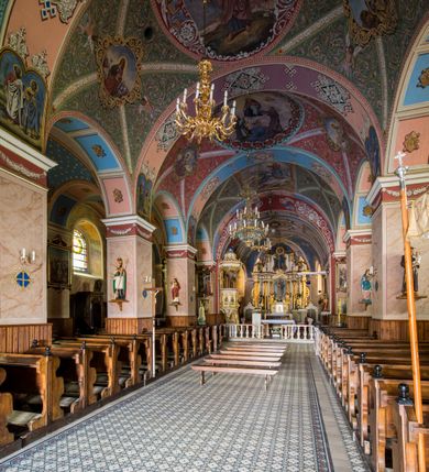 Zdjęcie nr 22: Kościół, zwrócony w kierunku południowo-wschodnim, jest budowlą murowaną z cegły i kamienia, na rzucie podłużnym. Długość bryły wynosi 35, a szerokość 16 metrów. Korpus nawowy jest trójnawowy i czteroprzęsłowy. Stosunkowo szerokie, jednoprzęsłowe, zamknięte półkoliście prezbiterium flankują dwie przyległości, z których jedna jest zakrystią. Na osi kościoła, w przedłużeniu północnego przęsła jest trójkondygnacyjna wieża, ujęta po bokach piętrowymi lokalnościami, lekko wysunięta przed lico fasady. Od zachodu do korpusu przylega kaplica. Nawy przekryte są sklepieniami kolebkowymi wspartymi na filarach o prostokątnym przekroju oraz pilastrach. Przestrzeń prezbiterium od korpusu nawowego oddziela drewniana balustrada. Artykulację ścian na zewnątrz kościoła stanowią osadzone na niewysokim cokole pojedyncze, a w narożach pary lizen wspierających obiegający bryłę fryz, przerwany w partii wieży oraz gzyms. Do wnętrza prowadzą trzy otwory drzwiowe – jeden w elewacji północnej oraz dwa w północnej ścianie kaplicy bocznej. Dwuskrzydłowe drzwi w fasadzie ujęte są profilowanym portalem zamkniętym łukiem odcinkowym zwieńczonym krzyżem. Po bokach wejścia znajduje się para kropielnic. Powyżej otworu drzwiowego usytuowane jest duże okno w kształcie stojącego prostokąta zamkniętego półkoliście, górą ujęte gzymsem. Na wysokości drugiej kondygnacji znajduje się mniejsze okno, zamknięte półkoliście i ujęte prostym obramieniem z kluczem pośrodku oraz parą guttusów pod parapetem. Podobną formę mają cztery okna w ścianach trzeciej kondygnacji wieży, dodatkowo wpisane są one w prostokątne płyciny zamknięte rodzajem łuku kotarowego. W górnej partii wieży znajdują się cztery, okrągłe otwory okienne ujęte prostymi obramieniami zwieńczonymi kluczem. Wieżę wieńczy hełm z osadzoną na cebulastej kopule latarnią zwieńczoną drugą, mniejszą zakończoną krzyżem. Ściany korpusu nawowego i prezbiterium przeprute są prostokątnymi oknami zamkniętymi półkoliście, ujętymi prostym obramieniem z kluczem pośrodku. Okno na zakończeniu prezbiterium jest zamurowane. Okna wypełnione są witrażami, które w prezbiterium zawierają wizerunki Najświętszego Serca Matki Boskiej po prawej i Najświętszego Serca Jezusa po lewej stronie.  Cykl witraży znajduje się także w oknach korpusu, po prawej stronie są to kolejno przedstawienia Matki Boskiej Bolesnej, Boga Ojca, św. Karola Borromeusza i św. Wojciecha, natomiast po lewej stronie – Męża Boleści, Ducha Świętego, św. Jana z Dukli oraz św. Stanisława. Kształt okien korpusu i prezbiterium został powtórzony w kaplicy bocznej, natomiast otwory okienne usytuowane w południowych ścianach przyległości flankujących prezbiterium są w kształcie stojących prostokątów. Korpus nawowy przekryty jest dachem dwuspadowym, wykonanym z blachy. Na wysokości pierwszego przęsła umieszczona jest wieżyczka na sygnaturkę, także obita blachą. Kolorystyka tynków kościoła utrzymana jest w odcieniu jasnożółtym, cokół jest szary natomiast detal architektoniczny jest biały. Plac przykościelny, z wybrukowanymi alejkami i trawnikiem, otoczony jest ogrodzeniem z murowanych słupków i metalowych segmentów.                    