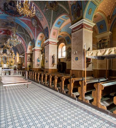 Zdjęcie nr 25: Kościół, zwrócony w kierunku południowo-wschodnim, jest budowlą murowaną z cegły i kamienia, na rzucie podłużnym. Długość bryły wynosi 35, a szerokość 16 metrów. Korpus nawowy jest trójnawowy i czteroprzęsłowy. Stosunkowo szerokie, jednoprzęsłowe, zamknięte półkoliście prezbiterium flankują dwie przyległości, z których jedna jest zakrystią. Na osi kościoła, w przedłużeniu północnego przęsła jest trójkondygnacyjna wieża, ujęta po bokach piętrowymi lokalnościami, lekko wysunięta przed lico fasady. Od zachodu do korpusu przylega kaplica. Nawy przekryte są sklepieniami kolebkowymi wspartymi na filarach o prostokątnym przekroju oraz pilastrach. Przestrzeń prezbiterium od korpusu nawowego oddziela drewniana balustrada. Artykulację ścian na zewnątrz kościoła stanowią osadzone na niewysokim cokole pojedyncze, a w narożach pary lizen wspierających obiegający bryłę fryz, przerwany w partii wieży oraz gzyms. Do wnętrza prowadzą trzy otwory drzwiowe – jeden w elewacji północnej oraz dwa w północnej ścianie kaplicy bocznej. Dwuskrzydłowe drzwi w fasadzie ujęte są profilowanym portalem zamkniętym łukiem odcinkowym zwieńczonym krzyżem. Po bokach wejścia znajduje się para kropielnic. Powyżej otworu drzwiowego usytuowane jest duże okno w kształcie stojącego prostokąta zamkniętego półkoliście, górą ujęte gzymsem. Na wysokości drugiej kondygnacji znajduje się mniejsze okno, zamknięte półkoliście i ujęte prostym obramieniem z kluczem pośrodku oraz parą guttusów pod parapetem. Podobną formę mają cztery okna w ścianach trzeciej kondygnacji wieży, dodatkowo wpisane są one w prostokątne płyciny zamknięte rodzajem łuku kotarowego. W górnej partii wieży znajdują się cztery, okrągłe otwory okienne ujęte prostymi obramieniami zwieńczonymi kluczem. Wieżę wieńczy hełm z osadzoną na cebulastej kopule latarnią zwieńczoną drugą, mniejszą zakończoną krzyżem. Ściany korpusu nawowego i prezbiterium przeprute są prostokątnymi oknami zamkniętymi półkoliście, ujętymi prostym obramieniem z kluczem pośrodku. Okno na zakończeniu prezbiterium jest zamurowane. Okna wypełnione są witrażami, które w prezbiterium zawierają wizerunki Najświętszego Serca Matki Boskiej po prawej i Najświętszego Serca Jezusa po lewej stronie.  Cykl witraży znajduje się także w oknach korpusu, po prawej stronie są to kolejno przedstawienia Matki Boskiej Bolesnej, Boga Ojca, św. Karola Borromeusza i św. Wojciecha, natomiast po lewej stronie – Męża Boleści, Ducha Świętego, św. Jana z Dukli oraz św. Stanisława. Kształt okien korpusu i prezbiterium został powtórzony w kaplicy bocznej, natomiast otwory okienne usytuowane w południowych ścianach przyległości flankujących prezbiterium są w kształcie stojących prostokątów. Korpus nawowy przekryty jest dachem dwuspadowym, wykonanym z blachy. Na wysokości pierwszego przęsła umieszczona jest wieżyczka na sygnaturkę, także obita blachą. Kolorystyka tynków kościoła utrzymana jest w odcieniu jasnożółtym, cokół jest szary natomiast detal architektoniczny jest biały. Plac przykościelny, z wybrukowanymi alejkami i trawnikiem, otoczony jest ogrodzeniem z murowanych słupków i metalowych segmentów.                    