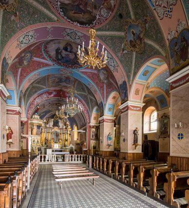 Zdjęcie nr 24: Kościół, zwrócony w kierunku południowo-wschodnim, jest budowlą murowaną z cegły i kamienia, na rzucie podłużnym. Długość bryły wynosi 35, a szerokość 16 metrów. Korpus nawowy jest trójnawowy i czteroprzęsłowy. Stosunkowo szerokie, jednoprzęsłowe, zamknięte półkoliście prezbiterium flankują dwie przyległości, z których jedna jest zakrystią. Na osi kościoła, w przedłużeniu północnego przęsła jest trójkondygnacyjna wieża, ujęta po bokach piętrowymi lokalnościami, lekko wysunięta przed lico fasady. Od zachodu do korpusu przylega kaplica. Nawy przekryte są sklepieniami kolebkowymi wspartymi na filarach o prostokątnym przekroju oraz pilastrach. Przestrzeń prezbiterium od korpusu nawowego oddziela drewniana balustrada. Artykulację ścian na zewnątrz kościoła stanowią osadzone na niewysokim cokole pojedyncze, a w narożach pary lizen wspierających obiegający bryłę fryz, przerwany w partii wieży oraz gzyms. Do wnętrza prowadzą trzy otwory drzwiowe – jeden w elewacji północnej oraz dwa w północnej ścianie kaplicy bocznej. Dwuskrzydłowe drzwi w fasadzie ujęte są profilowanym portalem zamkniętym łukiem odcinkowym zwieńczonym krzyżem. Po bokach wejścia znajduje się para kropielnic. Powyżej otworu drzwiowego usytuowane jest duże okno w kształcie stojącego prostokąta zamkniętego półkoliście, górą ujęte gzymsem. Na wysokości drugiej kondygnacji znajduje się mniejsze okno, zamknięte półkoliście i ujęte prostym obramieniem z kluczem pośrodku oraz parą guttusów pod parapetem. Podobną formę mają cztery okna w ścianach trzeciej kondygnacji wieży, dodatkowo wpisane są one w prostokątne płyciny zamknięte rodzajem łuku kotarowego. W górnej partii wieży znajdują się cztery, okrągłe otwory okienne ujęte prostymi obramieniami zwieńczonymi kluczem. Wieżę wieńczy hełm z osadzoną na cebulastej kopule latarnią zwieńczoną drugą, mniejszą zakończoną krzyżem. Ściany korpusu nawowego i prezbiterium przeprute są prostokątnymi oknami zamkniętymi półkoliście, ujętymi prostym obramieniem z kluczem pośrodku. Okno na zakończeniu prezbiterium jest zamurowane. Okna wypełnione są witrażami, które w prezbiterium zawierają wizerunki Najświętszego Serca Matki Boskiej po prawej i Najświętszego Serca Jezusa po lewej stronie.  Cykl witraży znajduje się także w oknach korpusu, po prawej stronie są to kolejno przedstawienia Matki Boskiej Bolesnej, Boga Ojca, św. Karola Borromeusza i św. Wojciecha, natomiast po lewej stronie – Męża Boleści, Ducha Świętego, św. Jana z Dukli oraz św. Stanisława. Kształt okien korpusu i prezbiterium został powtórzony w kaplicy bocznej, natomiast otwory okienne usytuowane w południowych ścianach przyległości flankujących prezbiterium są w kształcie stojących prostokątów. Korpus nawowy przekryty jest dachem dwuspadowym, wykonanym z blachy. Na wysokości pierwszego przęsła umieszczona jest wieżyczka na sygnaturkę, także obita blachą. Kolorystyka tynków kościoła utrzymana jest w odcieniu jasnożółtym, cokół jest szary natomiast detal architektoniczny jest biały. Plac przykościelny, z wybrukowanymi alejkami i trawnikiem, otoczony jest ogrodzeniem z murowanych słupków i metalowych segmentów.                    