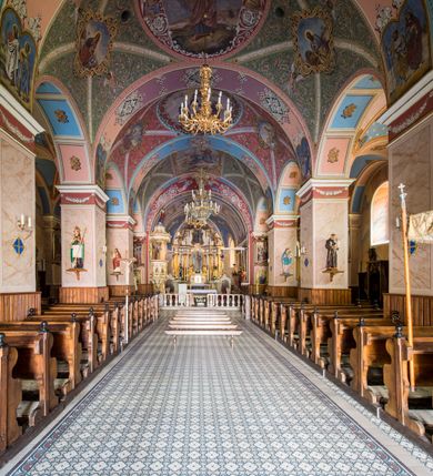 Zdjęcie nr 20: Kościół, zwrócony w kierunku południowo-wschodnim, jest budowlą murowaną z cegły i kamienia, na rzucie podłużnym. Długość bryły wynosi 35, a szerokość 16 metrów. Korpus nawowy jest trójnawowy i czteroprzęsłowy. Stosunkowo szerokie, jednoprzęsłowe, zamknięte półkoliście prezbiterium flankują dwie przyległości, z których jedna jest zakrystią. Na osi kościoła, w przedłużeniu północnego przęsła jest trójkondygnacyjna wieża, ujęta po bokach piętrowymi lokalnościami, lekko wysunięta przed lico fasady. Od zachodu do korpusu przylega kaplica. Nawy przekryte są sklepieniami kolebkowymi wspartymi na filarach o prostokątnym przekroju oraz pilastrach. Przestrzeń prezbiterium od korpusu nawowego oddziela drewniana balustrada. Artykulację ścian na zewnątrz kościoła stanowią osadzone na niewysokim cokole pojedyncze, a w narożach pary lizen wspierających obiegający bryłę fryz, przerwany w partii wieży oraz gzyms. Do wnętrza prowadzą trzy otwory drzwiowe – jeden w elewacji północnej oraz dwa w północnej ścianie kaplicy bocznej. Dwuskrzydłowe drzwi w fasadzie ujęte są profilowanym portalem zamkniętym łukiem odcinkowym zwieńczonym krzyżem. Po bokach wejścia znajduje się para kropielnic. Powyżej otworu drzwiowego usytuowane jest duże okno w kształcie stojącego prostokąta zamkniętego półkoliście, górą ujęte gzymsem. Na wysokości drugiej kondygnacji znajduje się mniejsze okno, zamknięte półkoliście i ujęte prostym obramieniem z kluczem pośrodku oraz parą guttusów pod parapetem. Podobną formę mają cztery okna w ścianach trzeciej kondygnacji wieży, dodatkowo wpisane są one w prostokątne płyciny zamknięte rodzajem łuku kotarowego. W górnej partii wieży znajdują się cztery, okrągłe otwory okienne ujęte prostymi obramieniami zwieńczonymi kluczem. Wieżę wieńczy hełm z osadzoną na cebulastej kopule latarnią zwieńczoną drugą, mniejszą zakończoną krzyżem. Ściany korpusu nawowego i prezbiterium przeprute są prostokątnymi oknami zamkniętymi półkoliście, ujętymi prostym obramieniem z kluczem pośrodku. Okno na zakończeniu prezbiterium jest zamurowane. Okna wypełnione są witrażami, które w prezbiterium zawierają wizerunki Najświętszego Serca Matki Boskiej po prawej i Najświętszego Serca Jezusa po lewej stronie.  Cykl witraży znajduje się także w oknach korpusu, po prawej stronie są to kolejno przedstawienia Matki Boskiej Bolesnej, Boga Ojca, św. Karola Borromeusza i św. Wojciecha, natomiast po lewej stronie – Męża Boleści, Ducha Świętego, św. Jana z Dukli oraz św. Stanisława. Kształt okien korpusu i prezbiterium został powtórzony w kaplicy bocznej, natomiast otwory okienne usytuowane w południowych ścianach przyległości flankujących prezbiterium są w kształcie stojących prostokątów. Korpus nawowy przekryty jest dachem dwuspadowym, wykonanym z blachy. Na wysokości pierwszego przęsła umieszczona jest wieżyczka na sygnaturkę, także obita blachą. Kolorystyka tynków kościoła utrzymana jest w odcieniu jasnożółtym, cokół jest szary natomiast detal architektoniczny jest biały. Plac przykościelny, z wybrukowanymi alejkami i trawnikiem, otoczony jest ogrodzeniem z murowanych słupków i metalowych segmentów.                    