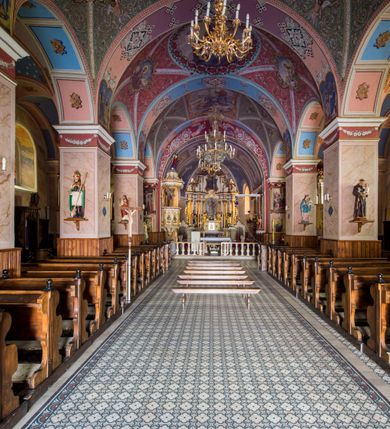 Zdjęcie nr 21: Kościół, zwrócony w kierunku południowo-wschodnim, jest budowlą murowaną z cegły i kamienia, na rzucie podłużnym. Długość bryły wynosi 35, a szerokość 16 metrów. Korpus nawowy jest trójnawowy i czteroprzęsłowy. Stosunkowo szerokie, jednoprzęsłowe, zamknięte półkoliście prezbiterium flankują dwie przyległości, z których jedna jest zakrystią. Na osi kościoła, w przedłużeniu północnego przęsła jest trójkondygnacyjna wieża, ujęta po bokach piętrowymi lokalnościami, lekko wysunięta przed lico fasady. Od zachodu do korpusu przylega kaplica. Nawy przekryte są sklepieniami kolebkowymi wspartymi na filarach o prostokątnym przekroju oraz pilastrach. Przestrzeń prezbiterium od korpusu nawowego oddziela drewniana balustrada. Artykulację ścian na zewnątrz kościoła stanowią osadzone na niewysokim cokole pojedyncze, a w narożach pary lizen wspierających obiegający bryłę fryz, przerwany w partii wieży oraz gzyms. Do wnętrza prowadzą trzy otwory drzwiowe – jeden w elewacji północnej oraz dwa w północnej ścianie kaplicy bocznej. Dwuskrzydłowe drzwi w fasadzie ujęte są profilowanym portalem zamkniętym łukiem odcinkowym zwieńczonym krzyżem. Po bokach wejścia znajduje się para kropielnic. Powyżej otworu drzwiowego usytuowane jest duże okno w kształcie stojącego prostokąta zamkniętego półkoliście, górą ujęte gzymsem. Na wysokości drugiej kondygnacji znajduje się mniejsze okno, zamknięte półkoliście i ujęte prostym obramieniem z kluczem pośrodku oraz parą guttusów pod parapetem. Podobną formę mają cztery okna w ścianach trzeciej kondygnacji wieży, dodatkowo wpisane są one w prostokątne płyciny zamknięte rodzajem łuku kotarowego. W górnej partii wieży znajdują się cztery, okrągłe otwory okienne ujęte prostymi obramieniami zwieńczonymi kluczem. Wieżę wieńczy hełm z osadzoną na cebulastej kopule latarnią zwieńczoną drugą, mniejszą zakończoną krzyżem. Ściany korpusu nawowego i prezbiterium przeprute są prostokątnymi oknami zamkniętymi półkoliście, ujętymi prostym obramieniem z kluczem pośrodku. Okno na zakończeniu prezbiterium jest zamurowane. Okna wypełnione są witrażami, które w prezbiterium zawierają wizerunki Najświętszego Serca Matki Boskiej po prawej i Najświętszego Serca Jezusa po lewej stronie.  Cykl witraży znajduje się także w oknach korpusu, po prawej stronie są to kolejno przedstawienia Matki Boskiej Bolesnej, Boga Ojca, św. Karola Borromeusza i św. Wojciecha, natomiast po lewej stronie – Męża Boleści, Ducha Świętego, św. Jana z Dukli oraz św. Stanisława. Kształt okien korpusu i prezbiterium został powtórzony w kaplicy bocznej, natomiast otwory okienne usytuowane w południowych ścianach przyległości flankujących prezbiterium są w kształcie stojących prostokątów. Korpus nawowy przekryty jest dachem dwuspadowym, wykonanym z blachy. Na wysokości pierwszego przęsła umieszczona jest wieżyczka na sygnaturkę, także obita blachą. Kolorystyka tynków kościoła utrzymana jest w odcieniu jasnożółtym, cokół jest szary natomiast detal architektoniczny jest biały. Plac przykościelny, z wybrukowanymi alejkami i trawnikiem, otoczony jest ogrodzeniem z murowanych słupków i metalowych segmentów.                    