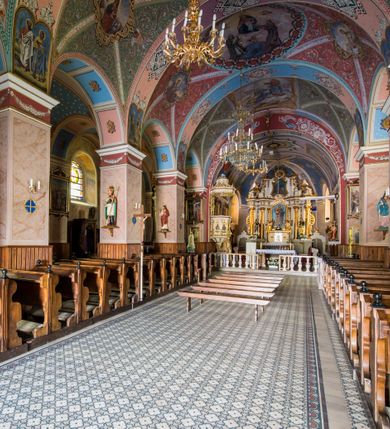 Zdjęcie nr 23: Kościół, zwrócony w kierunku południowo-wschodnim, jest budowlą murowaną z cegły i kamienia, na rzucie podłużnym. Długość bryły wynosi 35, a szerokość 16 metrów. Korpus nawowy jest trójnawowy i czteroprzęsłowy. Stosunkowo szerokie, jednoprzęsłowe, zamknięte półkoliście prezbiterium flankują dwie przyległości, z których jedna jest zakrystią. Na osi kościoła, w przedłużeniu północnego przęsła jest trójkondygnacyjna wieża, ujęta po bokach piętrowymi lokalnościami, lekko wysunięta przed lico fasady. Od zachodu do korpusu przylega kaplica. Nawy przekryte są sklepieniami kolebkowymi wspartymi na filarach o prostokątnym przekroju oraz pilastrach. Przestrzeń prezbiterium od korpusu nawowego oddziela drewniana balustrada. Artykulację ścian na zewnątrz kościoła stanowią osadzone na niewysokim cokole pojedyncze, a w narożach pary lizen wspierających obiegający bryłę fryz, przerwany w partii wieży oraz gzyms. Do wnętrza prowadzą trzy otwory drzwiowe – jeden w elewacji północnej oraz dwa w północnej ścianie kaplicy bocznej. Dwuskrzydłowe drzwi w fasadzie ujęte są profilowanym portalem zamkniętym łukiem odcinkowym zwieńczonym krzyżem. Po bokach wejścia znajduje się para kropielnic. Powyżej otworu drzwiowego usytuowane jest duże okno w kształcie stojącego prostokąta zamkniętego półkoliście, górą ujęte gzymsem. Na wysokości drugiej kondygnacji znajduje się mniejsze okno, zamknięte półkoliście i ujęte prostym obramieniem z kluczem pośrodku oraz parą guttusów pod parapetem. Podobną formę mają cztery okna w ścianach trzeciej kondygnacji wieży, dodatkowo wpisane są one w prostokątne płyciny zamknięte rodzajem łuku kotarowego. W górnej partii wieży znajdują się cztery, okrągłe otwory okienne ujęte prostymi obramieniami zwieńczonymi kluczem. Wieżę wieńczy hełm z osadzoną na cebulastej kopule latarnią zwieńczoną drugą, mniejszą zakończoną krzyżem. Ściany korpusu nawowego i prezbiterium przeprute są prostokątnymi oknami zamkniętymi półkoliście, ujętymi prostym obramieniem z kluczem pośrodku. Okno na zakończeniu prezbiterium jest zamurowane. Okna wypełnione są witrażami, które w prezbiterium zawierają wizerunki Najświętszego Serca Matki Boskiej po prawej i Najświętszego Serca Jezusa po lewej stronie.  Cykl witraży znajduje się także w oknach korpusu, po prawej stronie są to kolejno przedstawienia Matki Boskiej Bolesnej, Boga Ojca, św. Karola Borromeusza i św. Wojciecha, natomiast po lewej stronie – Męża Boleści, Ducha Świętego, św. Jana z Dukli oraz św. Stanisława. Kształt okien korpusu i prezbiterium został powtórzony w kaplicy bocznej, natomiast otwory okienne usytuowane w południowych ścianach przyległości flankujących prezbiterium są w kształcie stojących prostokątów. Korpus nawowy przekryty jest dachem dwuspadowym, wykonanym z blachy. Na wysokości pierwszego przęsła umieszczona jest wieżyczka na sygnaturkę, także obita blachą. Kolorystyka tynków kościoła utrzymana jest w odcieniu jasnożółtym, cokół jest szary natomiast detal architektoniczny jest biały. Plac przykościelny, z wybrukowanymi alejkami i trawnikiem, otoczony jest ogrodzeniem z murowanych słupków i metalowych segmentów.                    