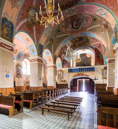 Zdjęcie nr 27: Kościół, zwrócony w kierunku południowo-wschodnim, jest budowlą murowaną z cegły i kamienia, na rzucie podłużnym. Długość bryły wynosi 35, a szerokość 16 metrów. Korpus nawowy jest trójnawowy i czteroprzęsłowy. Stosunkowo szerokie, jednoprzęsłowe, zamknięte półkoliście prezbiterium flankują dwie przyległości, z których jedna jest zakrystią. Na osi kościoła, w przedłużeniu północnego przęsła jest trójkondygnacyjna wieża, ujęta po bokach piętrowymi lokalnościami, lekko wysunięta przed lico fasady. Od zachodu do korpusu przylega kaplica. Nawy przekryte są sklepieniami kolebkowymi wspartymi na filarach o prostokątnym przekroju oraz pilastrach. Przestrzeń prezbiterium od korpusu nawowego oddziela drewniana balustrada. Artykulację ścian na zewnątrz kościoła stanowią osadzone na niewysokim cokole pojedyncze, a w narożach pary lizen wspierających obiegający bryłę fryz, przerwany w partii wieży oraz gzyms. Do wnętrza prowadzą trzy otwory drzwiowe – jeden w elewacji północnej oraz dwa w północnej ścianie kaplicy bocznej. Dwuskrzydłowe drzwi w fasadzie ujęte są profilowanym portalem zamkniętym łukiem odcinkowym zwieńczonym krzyżem. Po bokach wejścia znajduje się para kropielnic. Powyżej otworu drzwiowego usytuowane jest duże okno w kształcie stojącego prostokąta zamkniętego półkoliście, górą ujęte gzymsem. Na wysokości drugiej kondygnacji znajduje się mniejsze okno, zamknięte półkoliście i ujęte prostym obramieniem z kluczem pośrodku oraz parą guttusów pod parapetem. Podobną formę mają cztery okna w ścianach trzeciej kondygnacji wieży, dodatkowo wpisane są one w prostokątne płyciny zamknięte rodzajem łuku kotarowego. W górnej partii wieży znajdują się cztery, okrągłe otwory okienne ujęte prostymi obramieniami zwieńczonymi kluczem. Wieżę wieńczy hełm z osadzoną na cebulastej kopule latarnią zwieńczoną drugą, mniejszą zakończoną krzyżem. Ściany korpusu nawowego i prezbiterium przeprute są prostokątnymi oknami zamkniętymi półkoliście, ujętymi prostym obramieniem z kluczem pośrodku. Okno na zakończeniu prezbiterium jest zamurowane. Okna wypełnione są witrażami, które w prezbiterium zawierają wizerunki Najświętszego Serca Matki Boskiej po prawej i Najświętszego Serca Jezusa po lewej stronie.  Cykl witraży znajduje się także w oknach korpusu, po prawej stronie są to kolejno przedstawienia Matki Boskiej Bolesnej, Boga Ojca, św. Karola Borromeusza i św. Wojciecha, natomiast po lewej stronie – Męża Boleści, Ducha Świętego, św. Jana z Dukli oraz św. Stanisława. Kształt okien korpusu i prezbiterium został powtórzony w kaplicy bocznej, natomiast otwory okienne usytuowane w południowych ścianach przyległości flankujących prezbiterium są w kształcie stojących prostokątów. Korpus nawowy przekryty jest dachem dwuspadowym, wykonanym z blachy. Na wysokości pierwszego przęsła umieszczona jest wieżyczka na sygnaturkę, także obita blachą. Kolorystyka tynków kościoła utrzymana jest w odcieniu jasnożółtym, cokół jest szary natomiast detal architektoniczny jest biały. Plac przykościelny, z wybrukowanymi alejkami i trawnikiem, otoczony jest ogrodzeniem z murowanych słupków i metalowych segmentów.                    