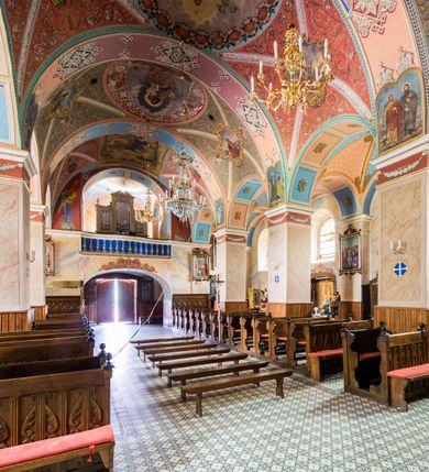 Zdjęcie nr 26: Kościół, zwrócony w kierunku południowo-wschodnim, jest budowlą murowaną z cegły i kamienia, na rzucie podłużnym. Długość bryły wynosi 35, a szerokość 16 metrów. Korpus nawowy jest trójnawowy i czteroprzęsłowy. Stosunkowo szerokie, jednoprzęsłowe, zamknięte półkoliście prezbiterium flankują dwie przyległości, z których jedna jest zakrystią. Na osi kościoła, w przedłużeniu północnego przęsła jest trójkondygnacyjna wieża, ujęta po bokach piętrowymi lokalnościami, lekko wysunięta przed lico fasady. Od zachodu do korpusu przylega kaplica. Nawy przekryte są sklepieniami kolebkowymi wspartymi na filarach o prostokątnym przekroju oraz pilastrach. Przestrzeń prezbiterium od korpusu nawowego oddziela drewniana balustrada. Artykulację ścian na zewnątrz kościoła stanowią osadzone na niewysokim cokole pojedyncze, a w narożach pary lizen wspierających obiegający bryłę fryz, przerwany w partii wieży oraz gzyms. Do wnętrza prowadzą trzy otwory drzwiowe – jeden w elewacji północnej oraz dwa w północnej ścianie kaplicy bocznej. Dwuskrzydłowe drzwi w fasadzie ujęte są profilowanym portalem zamkniętym łukiem odcinkowym zwieńczonym krzyżem. Po bokach wejścia znajduje się para kropielnic. Powyżej otworu drzwiowego usytuowane jest duże okno w kształcie stojącego prostokąta zamkniętego półkoliście, górą ujęte gzymsem. Na wysokości drugiej kondygnacji znajduje się mniejsze okno, zamknięte półkoliście i ujęte prostym obramieniem z kluczem pośrodku oraz parą guttusów pod parapetem. Podobną formę mają cztery okna w ścianach trzeciej kondygnacji wieży, dodatkowo wpisane są one w prostokątne płyciny zamknięte rodzajem łuku kotarowego. W górnej partii wieży znajdują się cztery, okrągłe otwory okienne ujęte prostymi obramieniami zwieńczonymi kluczem. Wieżę wieńczy hełm z osadzoną na cebulastej kopule latarnią zwieńczoną drugą, mniejszą zakończoną krzyżem. Ściany korpusu nawowego i prezbiterium przeprute są prostokątnymi oknami zamkniętymi półkoliście, ujętymi prostym obramieniem z kluczem pośrodku. Okno na zakończeniu prezbiterium jest zamurowane. Okna wypełnione są witrażami, które w prezbiterium zawierają wizerunki Najświętszego Serca Matki Boskiej po prawej i Najświętszego Serca Jezusa po lewej stronie.  Cykl witraży znajduje się także w oknach korpusu, po prawej stronie są to kolejno przedstawienia Matki Boskiej Bolesnej, Boga Ojca, św. Karola Borromeusza i św. Wojciecha, natomiast po lewej stronie – Męża Boleści, Ducha Świętego, św. Jana z Dukli oraz św. Stanisława. Kształt okien korpusu i prezbiterium został powtórzony w kaplicy bocznej, natomiast otwory okienne usytuowane w południowych ścianach przyległości flankujących prezbiterium są w kształcie stojących prostokątów. Korpus nawowy przekryty jest dachem dwuspadowym, wykonanym z blachy. Na wysokości pierwszego przęsła umieszczona jest wieżyczka na sygnaturkę, także obita blachą. Kolorystyka tynków kościoła utrzymana jest w odcieniu jasnożółtym, cokół jest szary natomiast detal architektoniczny jest biały. Plac przykościelny, z wybrukowanymi alejkami i trawnikiem, otoczony jest ogrodzeniem z murowanych słupków i metalowych segmentów.                    