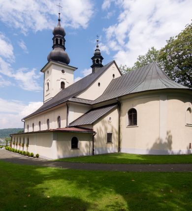 Zdjęcie nr 14: Kościół, zwrócony w kierunku południowo-wschodnim, jest budowlą murowaną z cegły i kamienia, na rzucie podłużnym. Długość bryły wynosi 35, a szerokość 16 metrów. Korpus nawowy jest trójnawowy i czteroprzęsłowy. Stosunkowo szerokie, jednoprzęsłowe, zamknięte półkoliście prezbiterium flankują dwie przyległości, z których jedna jest zakrystią. Na osi kościoła, w przedłużeniu północnego przęsła jest trójkondygnacyjna wieża, ujęta po bokach piętrowymi lokalnościami, lekko wysunięta przed lico fasady. Od zachodu do korpusu przylega kaplica. Nawy przekryte są sklepieniami kolebkowymi wspartymi na filarach o prostokątnym przekroju oraz pilastrach. Przestrzeń prezbiterium od korpusu nawowego oddziela drewniana balustrada. Artykulację ścian na zewnątrz kościoła stanowią osadzone na niewysokim cokole pojedyncze, a w narożach pary lizen wspierających obiegający bryłę fryz, przerwany w partii wieży oraz gzyms. Do wnętrza prowadzą trzy otwory drzwiowe – jeden w elewacji północnej oraz dwa w północnej ścianie kaplicy bocznej. Dwuskrzydłowe drzwi w fasadzie ujęte są profilowanym portalem zamkniętym łukiem odcinkowym zwieńczonym krzyżem. Po bokach wejścia znajduje się para kropielnic. Powyżej otworu drzwiowego usytuowane jest duże okno w kształcie stojącego prostokąta zamkniętego półkoliście, górą ujęte gzymsem. Na wysokości drugiej kondygnacji znajduje się mniejsze okno, zamknięte półkoliście i ujęte prostym obramieniem z kluczem pośrodku oraz parą guttusów pod parapetem. Podobną formę mają cztery okna w ścianach trzeciej kondygnacji wieży, dodatkowo wpisane są one w prostokątne płyciny zamknięte rodzajem łuku kotarowego. W górnej partii wieży znajdują się cztery, okrągłe otwory okienne ujęte prostymi obramieniami zwieńczonymi kluczem. Wieżę wieńczy hełm z osadzoną na cebulastej kopule latarnią zwieńczoną drugą, mniejszą zakończoną krzyżem. Ściany korpusu nawowego i prezbiterium przeprute są prostokątnymi oknami zamkniętymi półkoliście, ujętymi prostym obramieniem z kluczem pośrodku. Okno na zakończeniu prezbiterium jest zamurowane. Okna wypełnione są witrażami, które w prezbiterium zawierają wizerunki Najświętszego Serca Matki Boskiej po prawej i Najświętszego Serca Jezusa po lewej stronie.  Cykl witraży znajduje się także w oknach korpusu, po prawej stronie są to kolejno przedstawienia Matki Boskiej Bolesnej, Boga Ojca, św. Karola Borromeusza i św. Wojciecha, natomiast po lewej stronie – Męża Boleści, Ducha Świętego, św. Jana z Dukli oraz św. Stanisława. Kształt okien korpusu i prezbiterium został powtórzony w kaplicy bocznej, natomiast otwory okienne usytuowane w południowych ścianach przyległości flankujących prezbiterium są w kształcie stojących prostokątów. Korpus nawowy przekryty jest dachem dwuspadowym, wykonanym z blachy. Na wysokości pierwszego przęsła umieszczona jest wieżyczka na sygnaturkę, także obita blachą. Kolorystyka tynków kościoła utrzymana jest w odcieniu jasnożółtym, cokół jest szary natomiast detal architektoniczny jest biały. Plac przykościelny, z wybrukowanymi alejkami i trawnikiem, otoczony jest ogrodzeniem z murowanych słupków i metalowych segmentów.                    