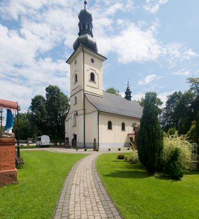 Zdjęcie nr 5: Kościół, zwrócony w kierunku południowo-wschodnim, jest budowlą murowaną z cegły i kamienia, na rzucie podłużnym. Długość bryły wynosi 35, a szerokość 16 metrów. Korpus nawowy jest trójnawowy i czteroprzęsłowy. Stosunkowo szerokie, jednoprzęsłowe, zamknięte półkoliście prezbiterium flankują dwie przyległości, z których jedna jest zakrystią. Na osi kościoła, w przedłużeniu północnego przęsła jest trójkondygnacyjna wieża, ujęta po bokach piętrowymi lokalnościami, lekko wysunięta przed lico fasady. Od zachodu do korpusu przylega kaplica. Nawy przekryte są sklepieniami kolebkowymi wspartymi na filarach o prostokątnym przekroju oraz pilastrach. Przestrzeń prezbiterium od korpusu nawowego oddziela drewniana balustrada. Artykulację ścian na zewnątrz kościoła stanowią osadzone na niewysokim cokole pojedyncze, a w narożach pary lizen wspierających obiegający bryłę fryz, przerwany w partii wieży oraz gzyms. Do wnętrza prowadzą trzy otwory drzwiowe – jeden w elewacji północnej oraz dwa w północnej ścianie kaplicy bocznej. Dwuskrzydłowe drzwi w fasadzie ujęte są profilowanym portalem zamkniętym łukiem odcinkowym zwieńczonym krzyżem. Po bokach wejścia znajduje się para kropielnic. Powyżej otworu drzwiowego usytuowane jest duże okno w kształcie stojącego prostokąta zamkniętego półkoliście, górą ujęte gzymsem. Na wysokości drugiej kondygnacji znajduje się mniejsze okno, zamknięte półkoliście i ujęte prostym obramieniem z kluczem pośrodku oraz parą guttusów pod parapetem. Podobną formę mają cztery okna w ścianach trzeciej kondygnacji wieży, dodatkowo wpisane są one w prostokątne płyciny zamknięte rodzajem łuku kotarowego. W górnej partii wieży znajdują się cztery, okrągłe otwory okienne ujęte prostymi obramieniami zwieńczonymi kluczem. Wieżę wieńczy hełm z osadzoną na cebulastej kopule latarnią zwieńczoną drugą, mniejszą zakończoną krzyżem. Ściany korpusu nawowego i prezbiterium przeprute są prostokątnymi oknami zamkniętymi półkoliście, ujętymi prostym obramieniem z kluczem pośrodku. Okno na zakończeniu prezbiterium jest zamurowane. Okna wypełnione są witrażami, które w prezbiterium zawierają wizerunki Najświętszego Serca Matki Boskiej po prawej i Najświętszego Serca Jezusa po lewej stronie.  Cykl witraży znajduje się także w oknach korpusu, po prawej stronie są to kolejno przedstawienia Matki Boskiej Bolesnej, Boga Ojca, św. Karola Borromeusza i św. Wojciecha, natomiast po lewej stronie – Męża Boleści, Ducha Świętego, św. Jana z Dukli oraz św. Stanisława. Kształt okien korpusu i prezbiterium został powtórzony w kaplicy bocznej, natomiast otwory okienne usytuowane w południowych ścianach przyległości flankujących prezbiterium są w kształcie stojących prostokątów. Korpus nawowy przekryty jest dachem dwuspadowym, wykonanym z blachy. Na wysokości pierwszego przęsła umieszczona jest wieżyczka na sygnaturkę, także obita blachą. Kolorystyka tynków kościoła utrzymana jest w odcieniu jasnożółtym, cokół jest szary natomiast detal architektoniczny jest biały. Plac przykościelny, z wybrukowanymi alejkami i trawnikiem, otoczony jest ogrodzeniem z murowanych słupków i metalowych segmentów.                    