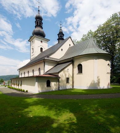 Zdjęcie nr 15: Kościół, zwrócony w kierunku południowo-wschodnim, jest budowlą murowaną z cegły i kamienia, na rzucie podłużnym. Długość bryły wynosi 35, a szerokość 16 metrów. Korpus nawowy jest trójnawowy i czteroprzęsłowy. Stosunkowo szerokie, jednoprzęsłowe, zamknięte półkoliście prezbiterium flankują dwie przyległości, z których jedna jest zakrystią. Na osi kościoła, w przedłużeniu północnego przęsła jest trójkondygnacyjna wieża, ujęta po bokach piętrowymi lokalnościami, lekko wysunięta przed lico fasady. Od zachodu do korpusu przylega kaplica. Nawy przekryte są sklepieniami kolebkowymi wspartymi na filarach o prostokątnym przekroju oraz pilastrach. Przestrzeń prezbiterium od korpusu nawowego oddziela drewniana balustrada. Artykulację ścian na zewnątrz kościoła stanowią osadzone na niewysokim cokole pojedyncze, a w narożach pary lizen wspierających obiegający bryłę fryz, przerwany w partii wieży oraz gzyms. Do wnętrza prowadzą trzy otwory drzwiowe – jeden w elewacji północnej oraz dwa w północnej ścianie kaplicy bocznej. Dwuskrzydłowe drzwi w fasadzie ujęte są profilowanym portalem zamkniętym łukiem odcinkowym zwieńczonym krzyżem. Po bokach wejścia znajduje się para kropielnic. Powyżej otworu drzwiowego usytuowane jest duże okno w kształcie stojącego prostokąta zamkniętego półkoliście, górą ujęte gzymsem. Na wysokości drugiej kondygnacji znajduje się mniejsze okno, zamknięte półkoliście i ujęte prostym obramieniem z kluczem pośrodku oraz parą guttusów pod parapetem. Podobną formę mają cztery okna w ścianach trzeciej kondygnacji wieży, dodatkowo wpisane są one w prostokątne płyciny zamknięte rodzajem łuku kotarowego. W górnej partii wieży znajdują się cztery, okrągłe otwory okienne ujęte prostymi obramieniami zwieńczonymi kluczem. Wieżę wieńczy hełm z osadzoną na cebulastej kopule latarnią zwieńczoną drugą, mniejszą zakończoną krzyżem. Ściany korpusu nawowego i prezbiterium przeprute są prostokątnymi oknami zamkniętymi półkoliście, ujętymi prostym obramieniem z kluczem pośrodku. Okno na zakończeniu prezbiterium jest zamurowane. Okna wypełnione są witrażami, które w prezbiterium zawierają wizerunki Najświętszego Serca Matki Boskiej po prawej i Najświętszego Serca Jezusa po lewej stronie.  Cykl witraży znajduje się także w oknach korpusu, po prawej stronie są to kolejno przedstawienia Matki Boskiej Bolesnej, Boga Ojca, św. Karola Borromeusza i św. Wojciecha, natomiast po lewej stronie – Męża Boleści, Ducha Świętego, św. Jana z Dukli oraz św. Stanisława. Kształt okien korpusu i prezbiterium został powtórzony w kaplicy bocznej, natomiast otwory okienne usytuowane w południowych ścianach przyległości flankujących prezbiterium są w kształcie stojących prostokątów. Korpus nawowy przekryty jest dachem dwuspadowym, wykonanym z blachy. Na wysokości pierwszego przęsła umieszczona jest wieżyczka na sygnaturkę, także obita blachą. Kolorystyka tynków kościoła utrzymana jest w odcieniu jasnożółtym, cokół jest szary natomiast detal architektoniczny jest biały. Plac przykościelny, z wybrukowanymi alejkami i trawnikiem, otoczony jest ogrodzeniem z murowanych słupków i metalowych segmentów.                    
