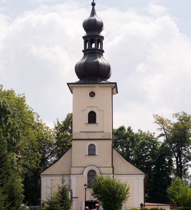 Zdjęcie nr 13: Kościół, zwrócony w kierunku południowo-wschodnim, jest budowlą murowaną z cegły i kamienia, na rzucie podłużnym. Długość bryły wynosi 35, a szerokość 16 metrów. Korpus nawowy jest trójnawowy i czteroprzęsłowy. Stosunkowo szerokie, jednoprzęsłowe, zamknięte półkoliście prezbiterium flankują dwie przyległości, z których jedna jest zakrystią. Na osi kościoła, w przedłużeniu północnego przęsła jest trójkondygnacyjna wieża, ujęta po bokach piętrowymi lokalnościami, lekko wysunięta przed lico fasady. Od zachodu do korpusu przylega kaplica. Nawy przekryte są sklepieniami kolebkowymi wspartymi na filarach o prostokątnym przekroju oraz pilastrach. Przestrzeń prezbiterium od korpusu nawowego oddziela drewniana balustrada. Artykulację ścian na zewnątrz kościoła stanowią osadzone na niewysokim cokole pojedyncze, a w narożach pary lizen wspierających obiegający bryłę fryz, przerwany w partii wieży oraz gzyms. Do wnętrza prowadzą trzy otwory drzwiowe – jeden w elewacji północnej oraz dwa w północnej ścianie kaplicy bocznej. Dwuskrzydłowe drzwi w fasadzie ujęte są profilowanym portalem zamkniętym łukiem odcinkowym zwieńczonym krzyżem. Po bokach wejścia znajduje się para kropielnic. Powyżej otworu drzwiowego usytuowane jest duże okno w kształcie stojącego prostokąta zamkniętego półkoliście, górą ujęte gzymsem. Na wysokości drugiej kondygnacji znajduje się mniejsze okno, zamknięte półkoliście i ujęte prostym obramieniem z kluczem pośrodku oraz parą guttusów pod parapetem. Podobną formę mają cztery okna w ścianach trzeciej kondygnacji wieży, dodatkowo wpisane są one w prostokątne płyciny zamknięte rodzajem łuku kotarowego. W górnej partii wieży znajdują się cztery, okrągłe otwory okienne ujęte prostymi obramieniami zwieńczonymi kluczem. Wieżę wieńczy hełm z osadzoną na cebulastej kopule latarnią zwieńczoną drugą, mniejszą zakończoną krzyżem. Ściany korpusu nawowego i prezbiterium przeprute są prostokątnymi oknami zamkniętymi półkoliście, ujętymi prostym obramieniem z kluczem pośrodku. Okno na zakończeniu prezbiterium jest zamurowane. Okna wypełnione są witrażami, które w prezbiterium zawierają wizerunki Najświętszego Serca Matki Boskiej po prawej i Najświętszego Serca Jezusa po lewej stronie.  Cykl witraży znajduje się także w oknach korpusu, po prawej stronie są to kolejno przedstawienia Matki Boskiej Bolesnej, Boga Ojca, św. Karola Borromeusza i św. Wojciecha, natomiast po lewej stronie – Męża Boleści, Ducha Świętego, św. Jana z Dukli oraz św. Stanisława. Kształt okien korpusu i prezbiterium został powtórzony w kaplicy bocznej, natomiast otwory okienne usytuowane w południowych ścianach przyległości flankujących prezbiterium są w kształcie stojących prostokątów. Korpus nawowy przekryty jest dachem dwuspadowym, wykonanym z blachy. Na wysokości pierwszego przęsła umieszczona jest wieżyczka na sygnaturkę, także obita blachą. Kolorystyka tynków kościoła utrzymana jest w odcieniu jasnożółtym, cokół jest szary natomiast detal architektoniczny jest biały. Plac przykościelny, z wybrukowanymi alejkami i trawnikiem, otoczony jest ogrodzeniem z murowanych słupków i metalowych segmentów.                    